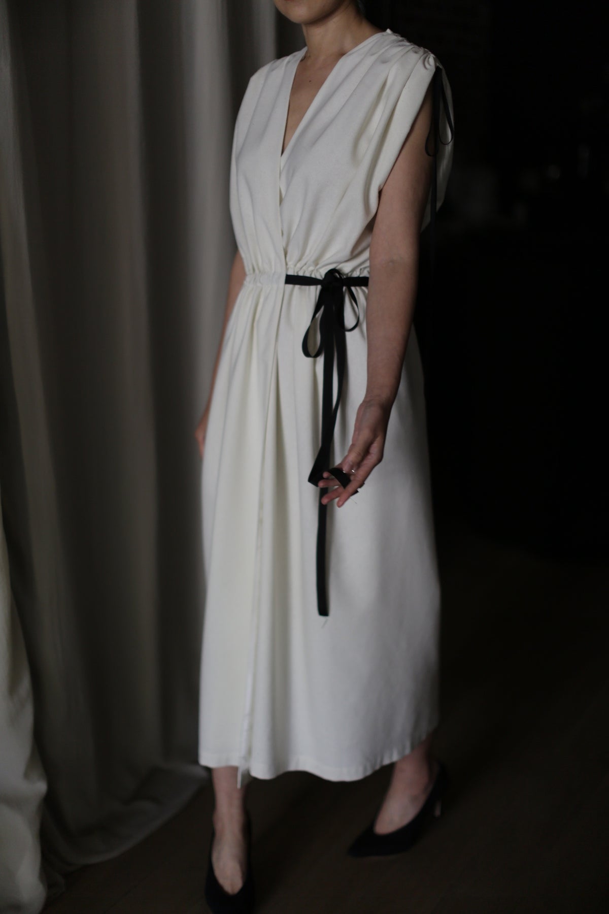 Person wearing a sleeveless, white raw silk Lune dress with a black ribbon tied at the waist. The background is dimly lit and appears to include curtains, and the person’s face is outside the frame. The person is also wearing black pointed-toe shoes.