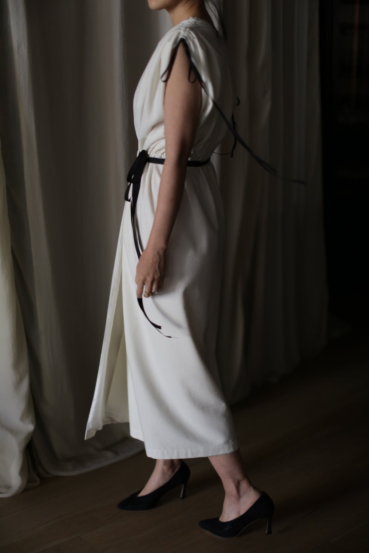 A person stands indoors wearing the Lune, a sleeveless, ankle-length handmade dress tied with a black belt. The back straps of the dress hang loosely as they are seen in profile, with black high heels on a wooden floor and cream-colored curtains in the background.