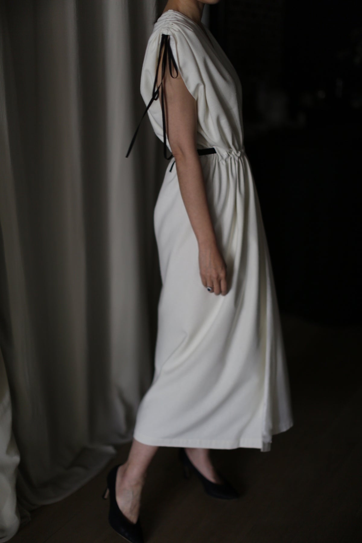 A person wearing the Lune, a flowing, sleeveless white raw silk dress with black ribbon detailing at the shoulders and around the waist, stands beside a curtain. Paired with black heels, this handmade dress encapsulates elegance in dim lighting, creating a soft and sophisticated atmosphere.