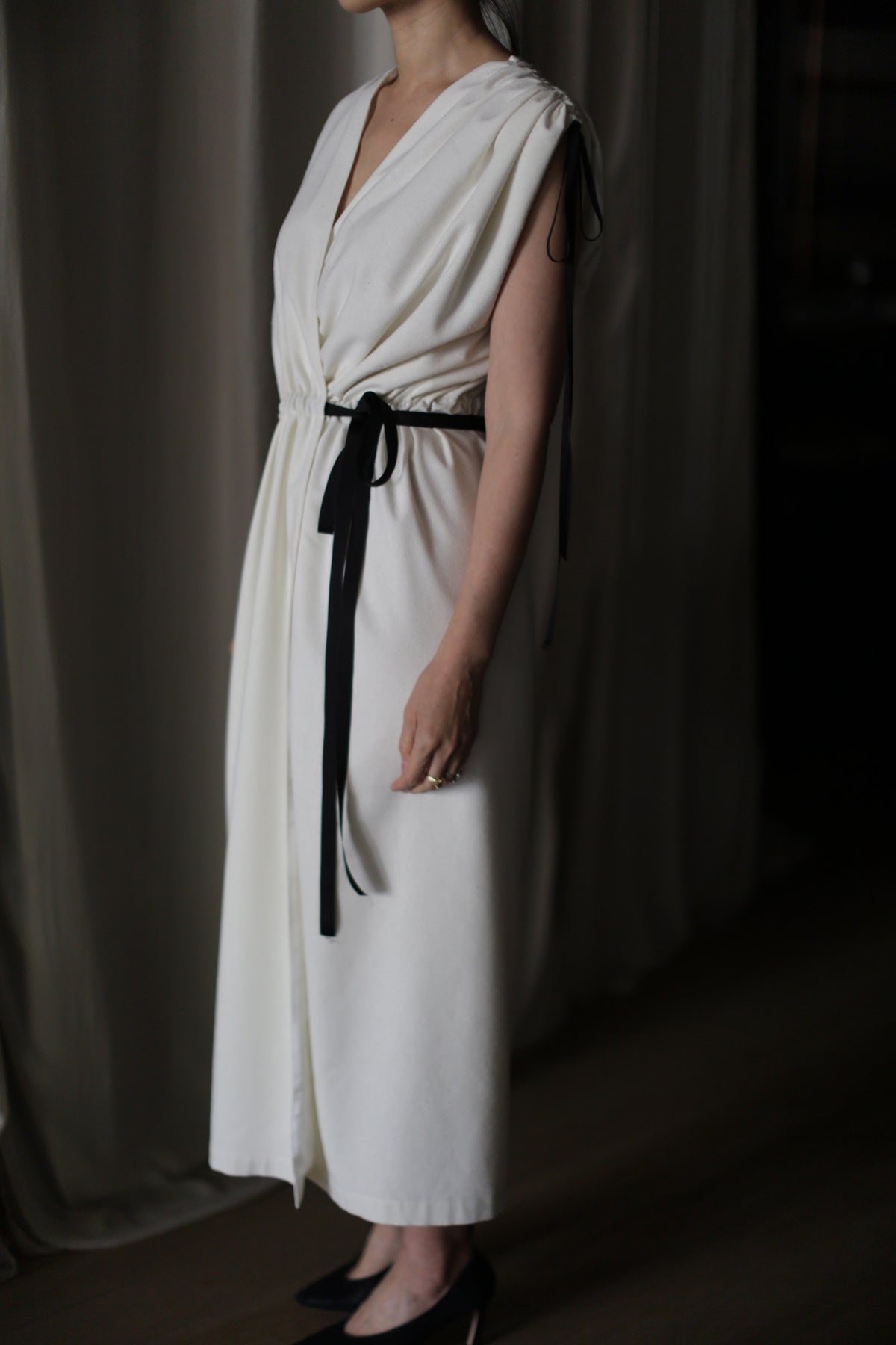 A person stands against a dark background, wearing the Lune dress—an elegant, handmade long dress crafted from white raw silk with a V-neckline and a black ribbon belt tied at the waist. Only the lower part of their face and part of their upper body are visible in the dim lighting, showcasing the sophistication of this sleeveless design.