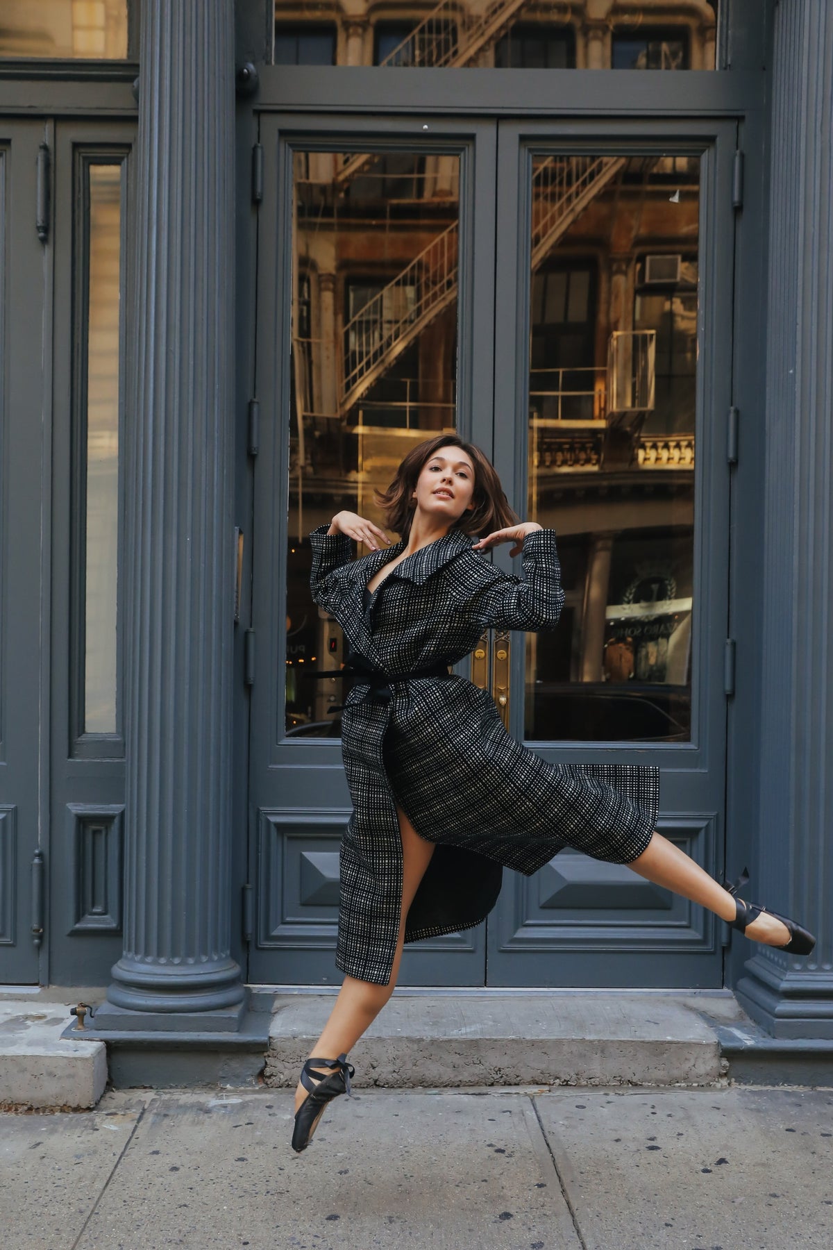 A woman in a luxurious Malvern double-breasted coat gracefully leaps in front of a blue door with large windows, reflecting a building adorned with intricate metal railings. She wears black shoes, and her movement adds a dynamic flair to the urban backdrop.