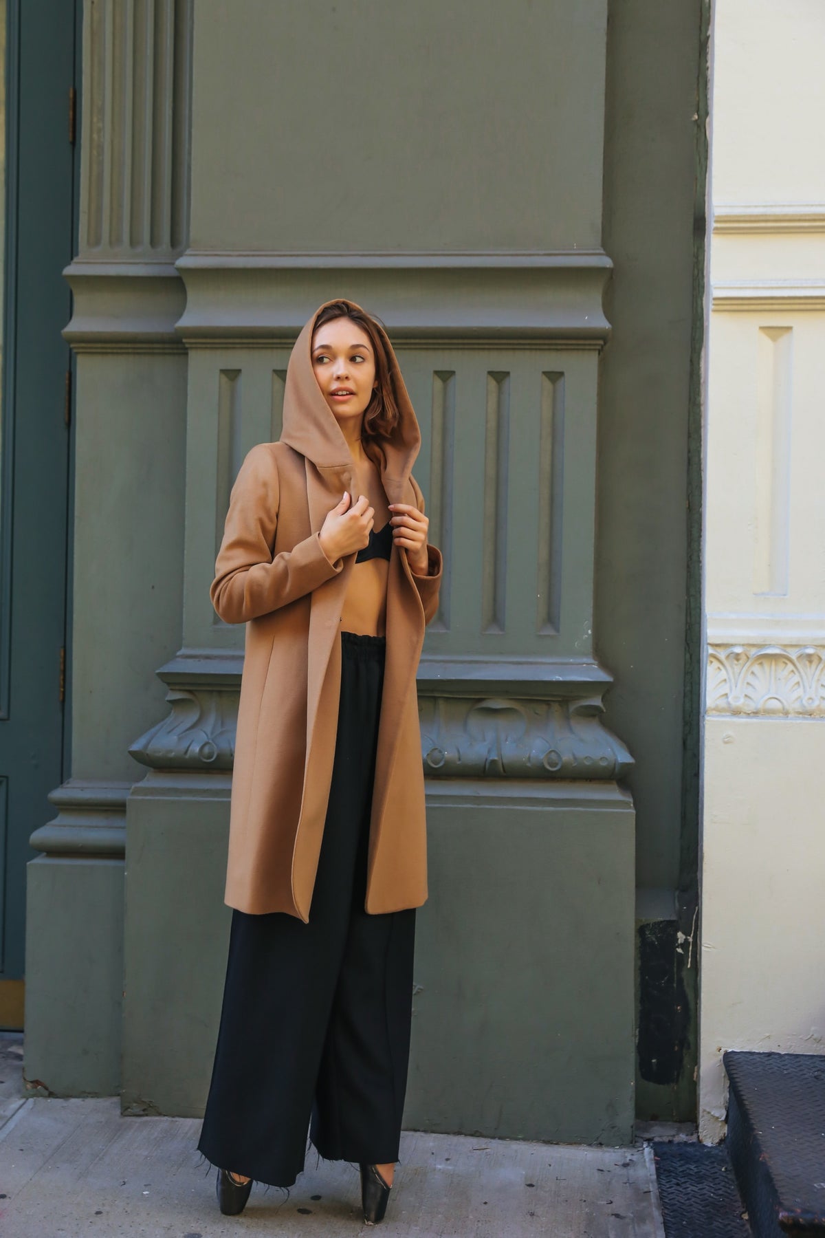 A woman in a fairytale-like Mitchley Long tan coat with a hood stands against an ornate building facade. Her woven cashmere ensemble pairs with black wide-leg trousers and heels, enhanced by handmade snap buttons, as she gazes thoughtfully to the side.