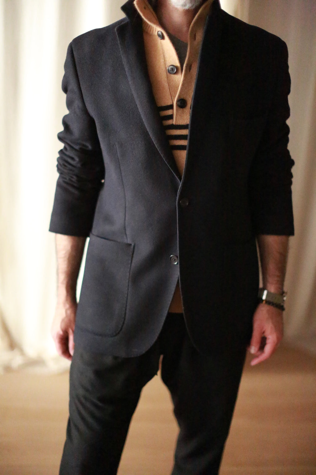 A person wearing a Men's Cashmere Blazer in a stylish black hue over a tan sweater with black stripes stands with hands partially visible. The softly lit, neutral background enhances the smart-casual attire, showcasing a refined European fit that exudes sophistication.