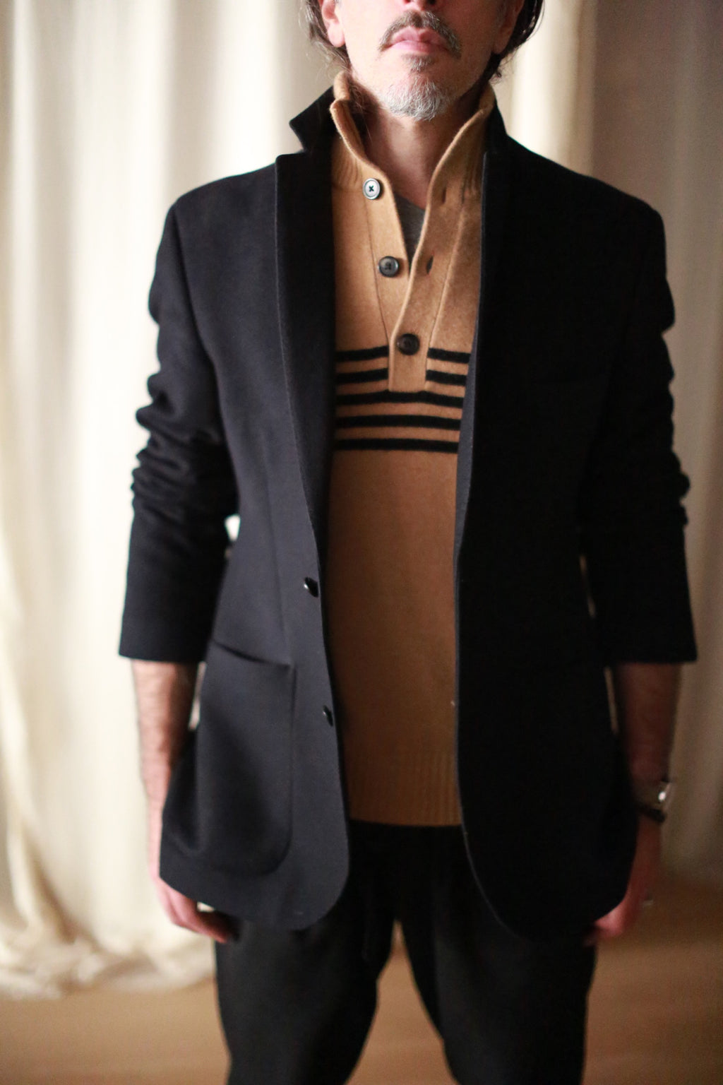 A person is wearing a Men's Cashmere Blazer in a European fit, complemented by a tan sweater featuring black horizontal stripes. The background is softly lit, and the individual stands with their face partially out of view.