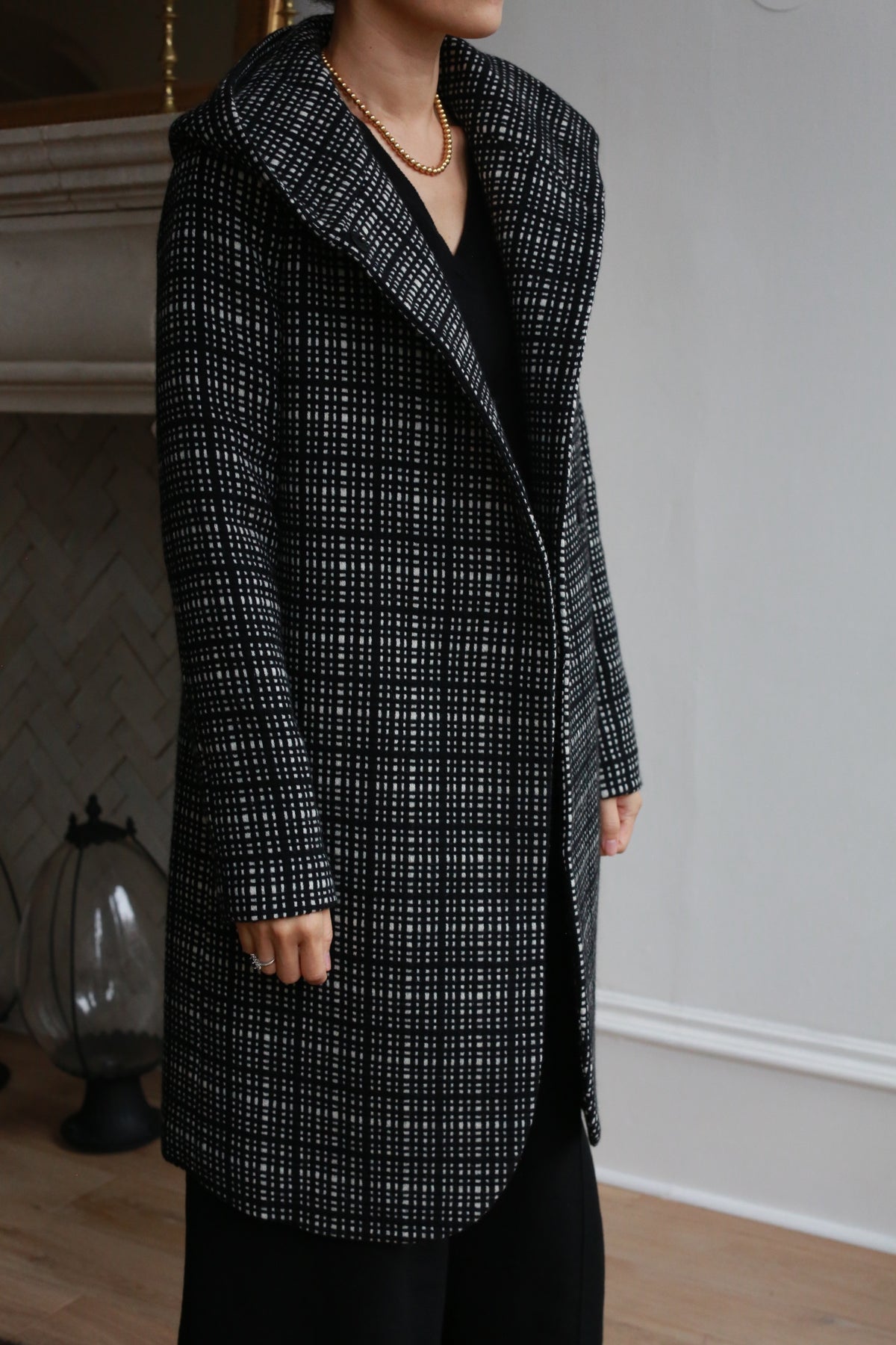 A person wearing the Mitchley Long | Plaid cashmere coat stands indoors by a fireplace, a gold chain necklace visible. Their head is partly out of frame, drawing attention to the elegant plaid design of the jacket.