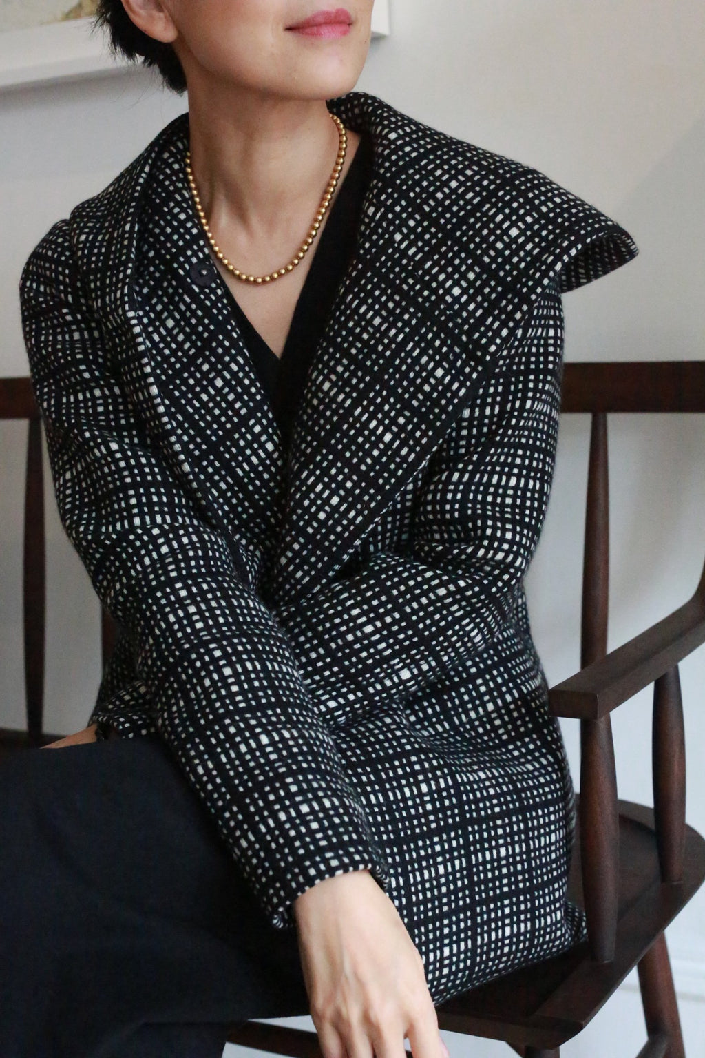 A woman with short dark hair, wearing the Mitchley | Plaid black and white checkered coat and a gold beaded necklace, sits on a wooden chair with her face partially visible.