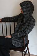 A person wearing the Mitchley | Plaid black and white hooded jacket sits sideways on a wooden bench, facing left, their face partially hidden by the hood.