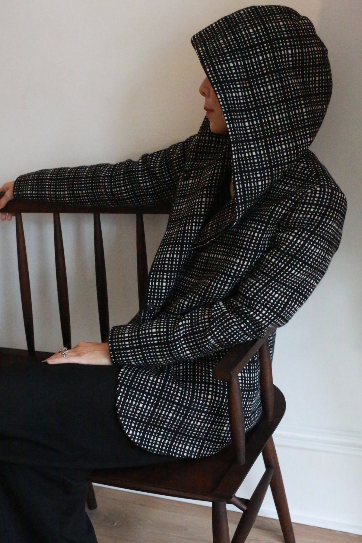 A person wearing the Mitchley | Plaid black and white hooded jacket sits sideways on a wooden bench, facing left, their face partially hidden by the hood.
