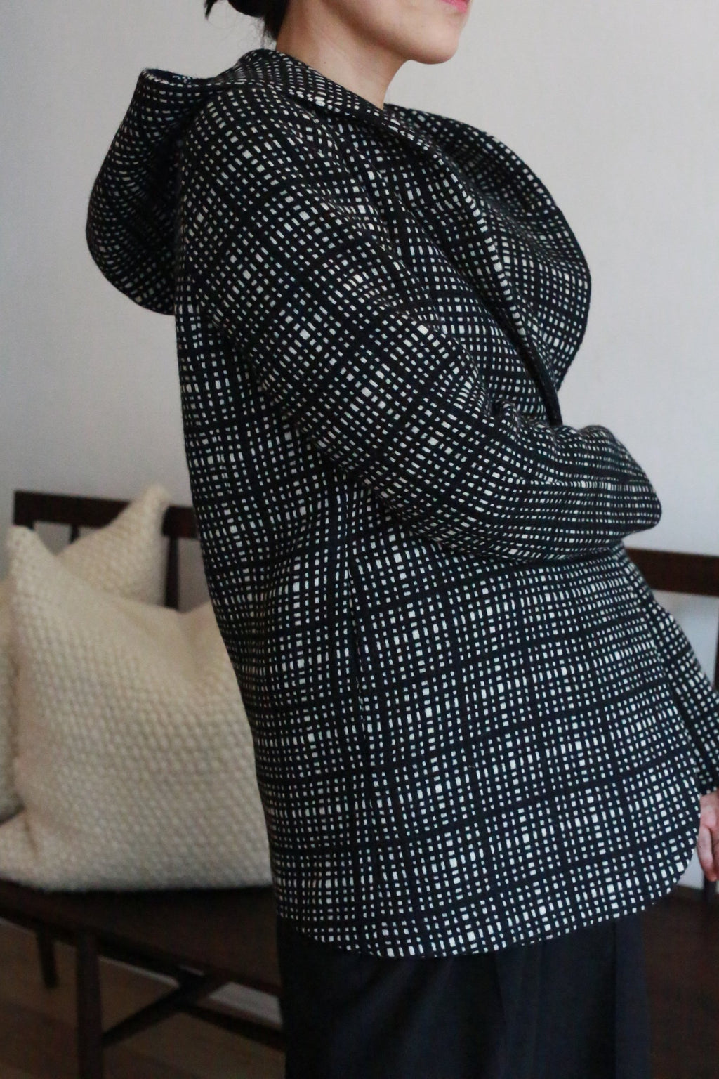 A person wearing the Mitchley | Plaid jacket stands sideways indoors, with white textured pillows and a wooden bench in the background.