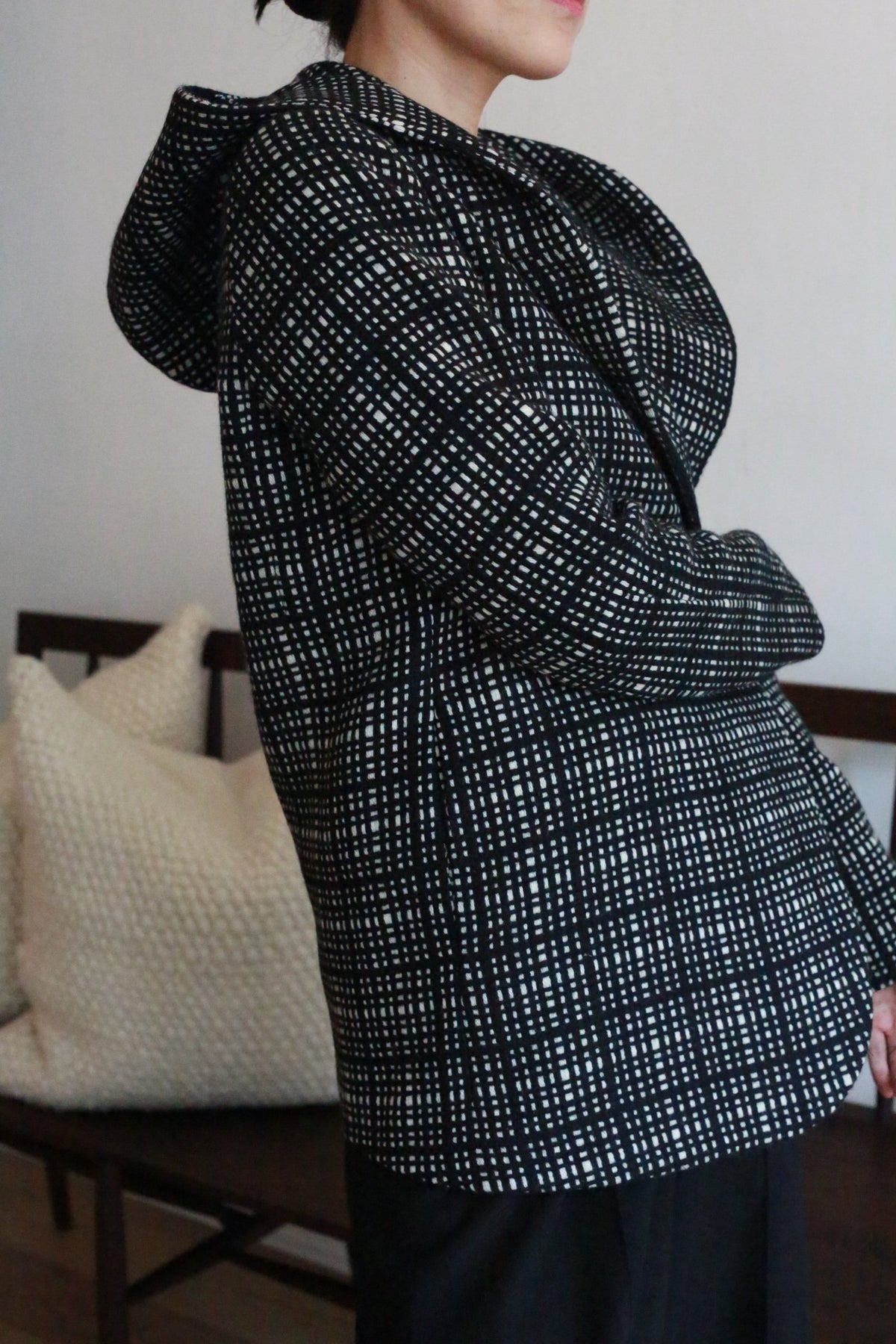 A person wearing the Mitchley | Plaid jacket stands sideways indoors, with white textured pillows and a wooden bench in the background.