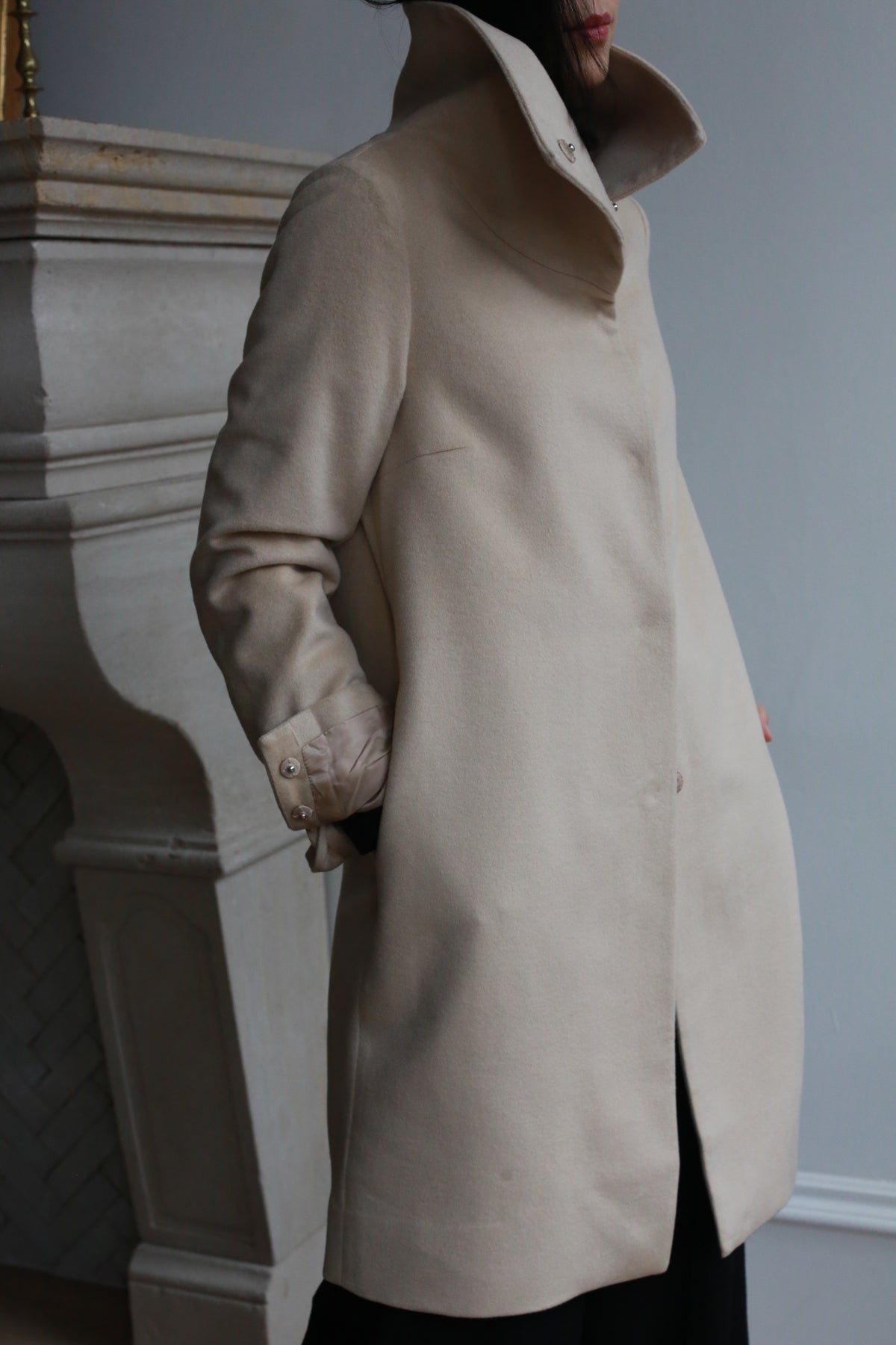 A person stands with their hand in the pocket of a Stamford Long coat, crafted from 100% woven cashmere. The dramatic collar adds flair against the backdrop of a decorative white wall.