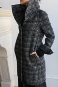 Indoors, a person wears the Stamford Long | Plaid coat in black and white cashmere, featuring a bold plaid pattern, large high collar, and pockets. Their face is partially turned away from the camera.