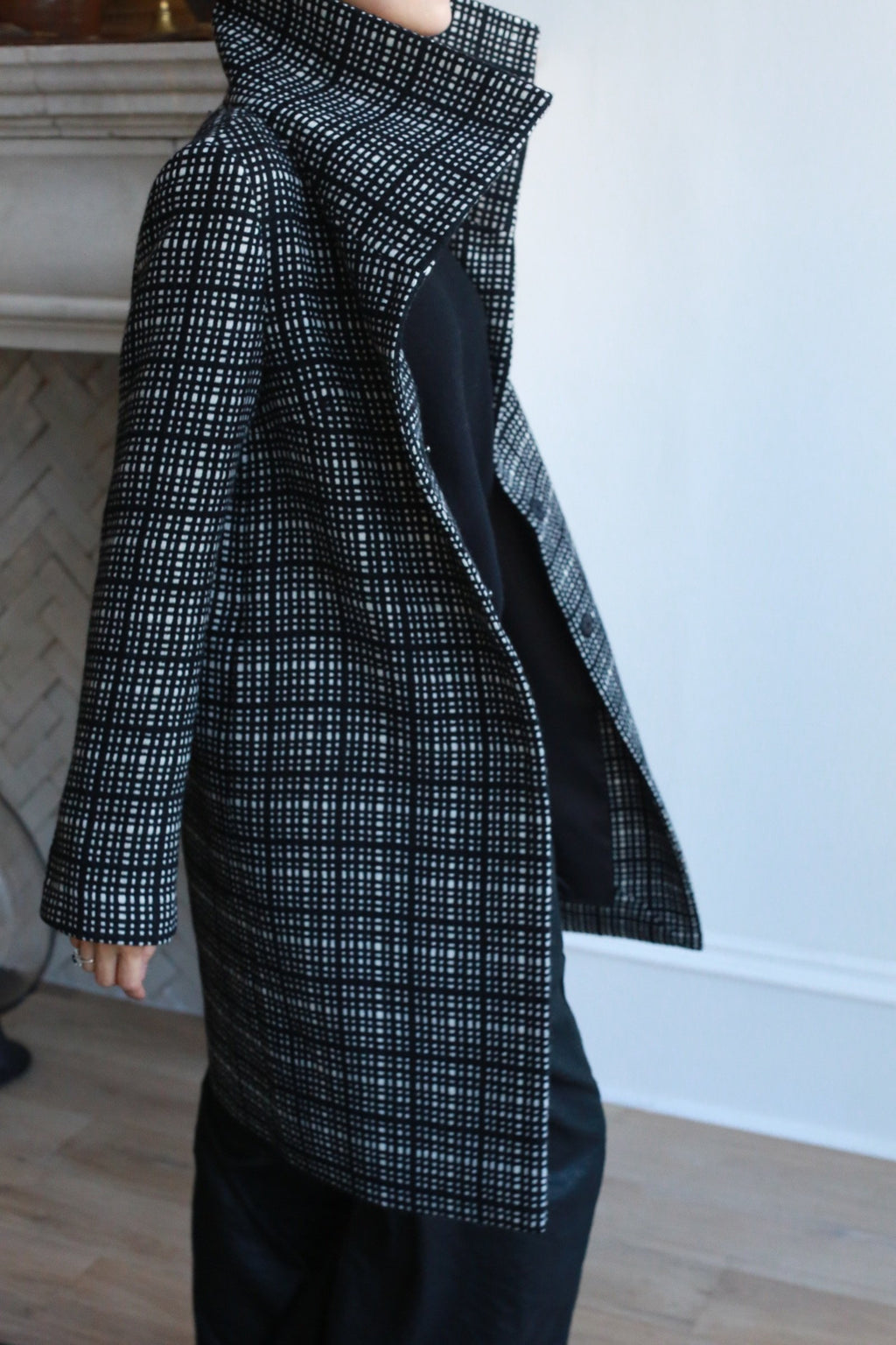 A person stands indoors by a light wall and fireplace, wearing the Stamford Long | Plaid coat with a high collar, paired with black pants and a black top. Their face is not visible.