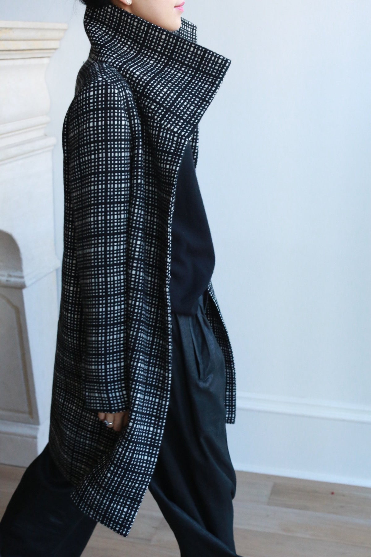 A person wearing the Stamford Long | Plaid coat in black and white cashmere with a high collar, black top, and loose black pants walks indoors against a light wall and wooden floor.