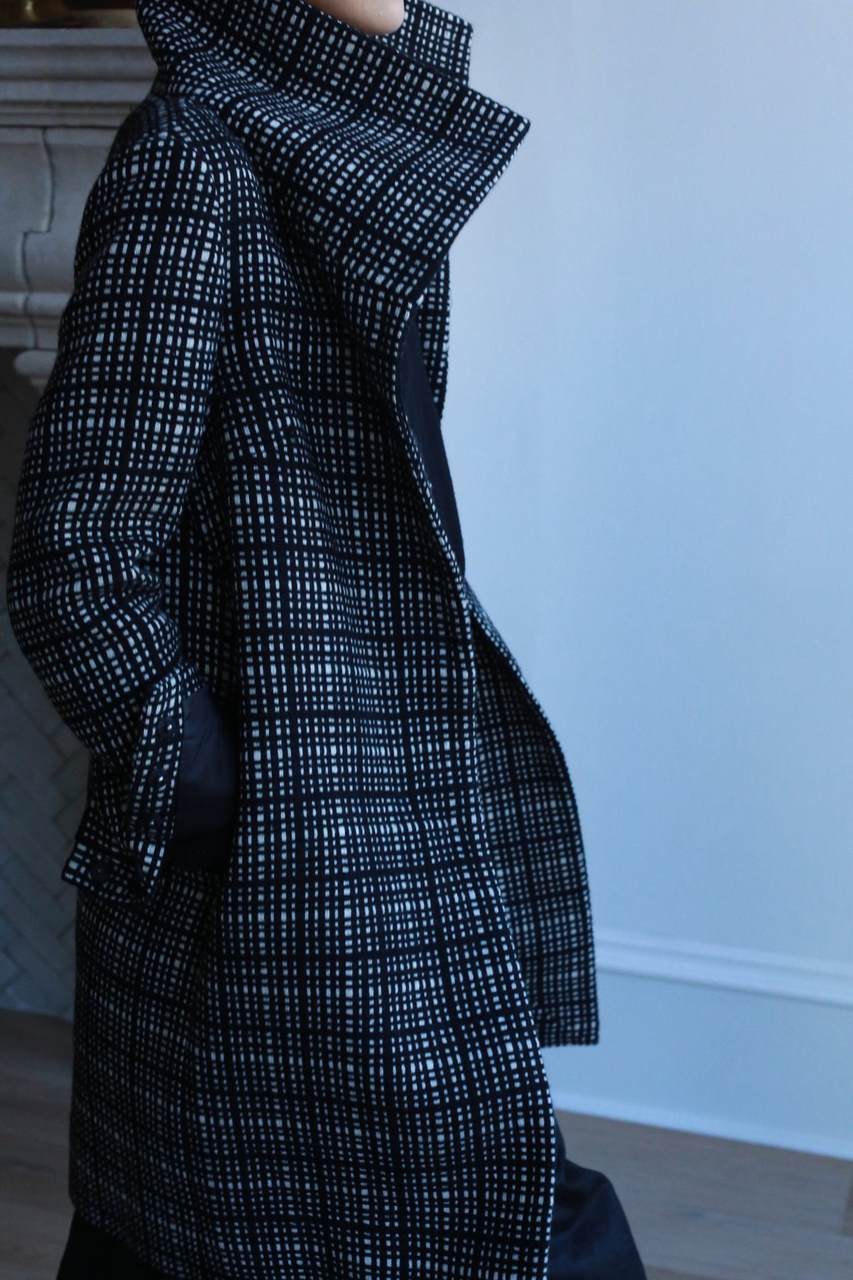 Someone stands indoors, hands in pockets, wearing the Stamford Long | Plaid coat in black and white cashmere with a high collar. The softly lit, minimal background enhances the refined appearance of both the coat and its wearer.