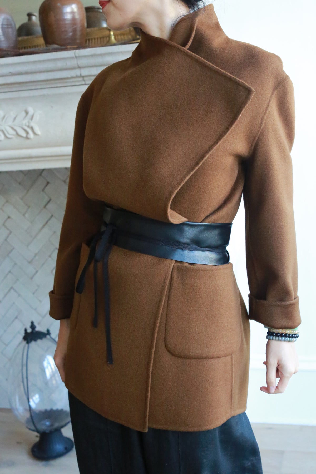 A person wearing a stylish brown coat crafted from genuine lambskin with an asymmetrical design and the Leather Obi with Grosgrain Ribbon around the waist stands indoors. The background features a decorative fireplace and vases. The person's face is not shown.
