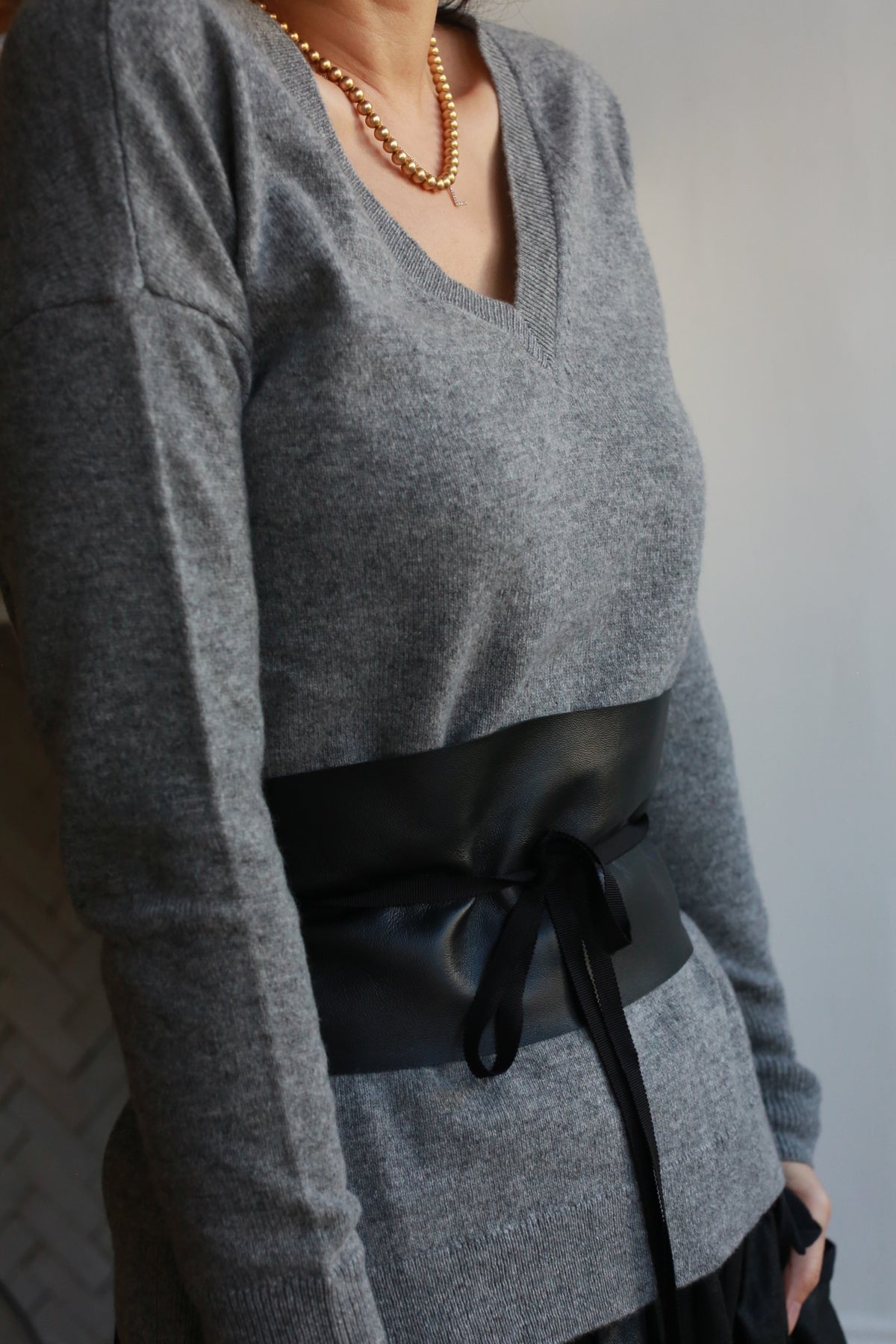 A person wears a gray V-neck sweater adorned with a gold chain necklace. A Leather Obi with Grosgrain Ribbon, made of genuine lambskin and featuring a tied bow, accentuates their waist. The background is a neutral-toned wall.