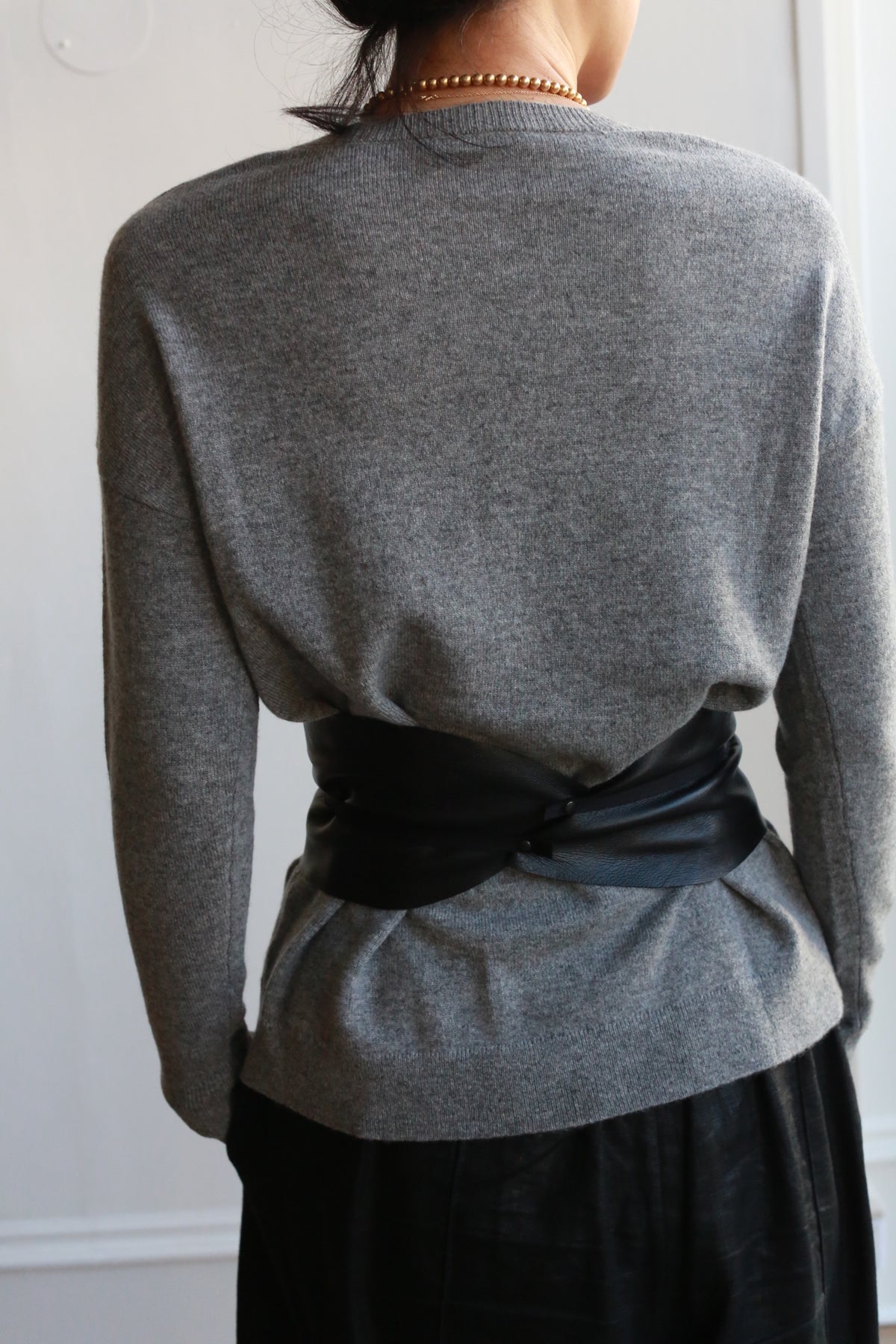 A person with long hair is seen from the back, wearing a gray sweater. They have a Leather Obi with Grosgrain Ribbon cinched around their waist, and a golden necklace is visible around their neck. The background is minimal and neutral.