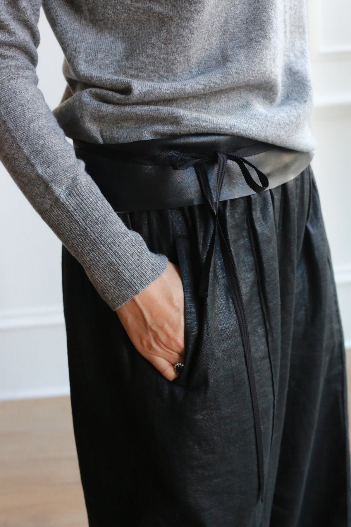 Close-up of a person wearing a gray knit sweater tucked into high-waisted, loose-fitting black pants. The pants are accentuated by the Leather Obi with Grosgrain Ribbon, stylishly tied in a bow at the front. A hand is casually placed in one pocket, highlighting this versatile accessory.