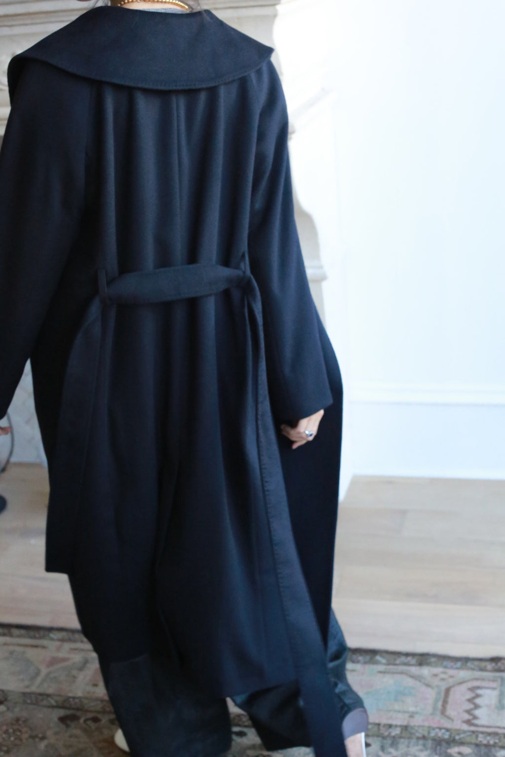 A person walks on a hardwood floor wearing the Malvern, a long, double-breasted luxury coat featuring a wide collar and belt. They are in a room with a light-colored wall, where part of a patterned rug is visible. The image captures the scene from behind.