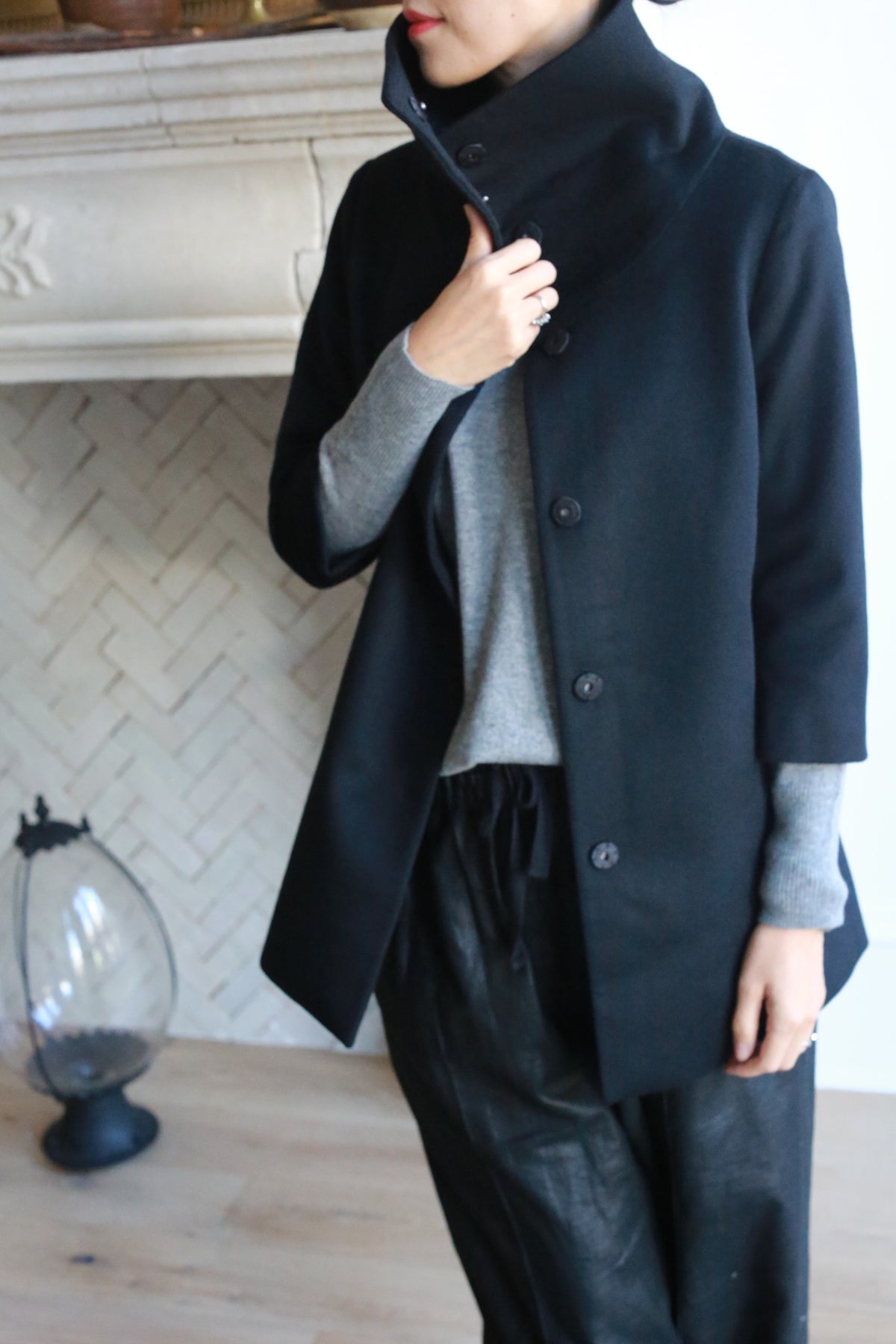 A person stands indoors wearing the black high-collared Stamford jacket, a gray cashmere long-sleeve shirt, and black pants. Their left hand is touching the collar. They are near a white brick fireplace, with a decorative item partially visible on the floor to the left.