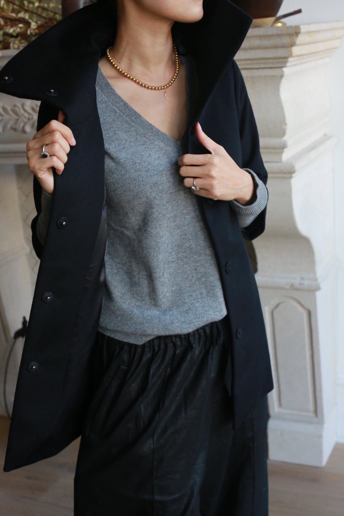 A woman is seen from the shoulders down wearing the Stamford, a dark coat with an upturned high collar, paired with a gray cashmere V-neck sweater and black pants. She accessorizes with a gold necklace and rings, standing in front of a white architectural feature.