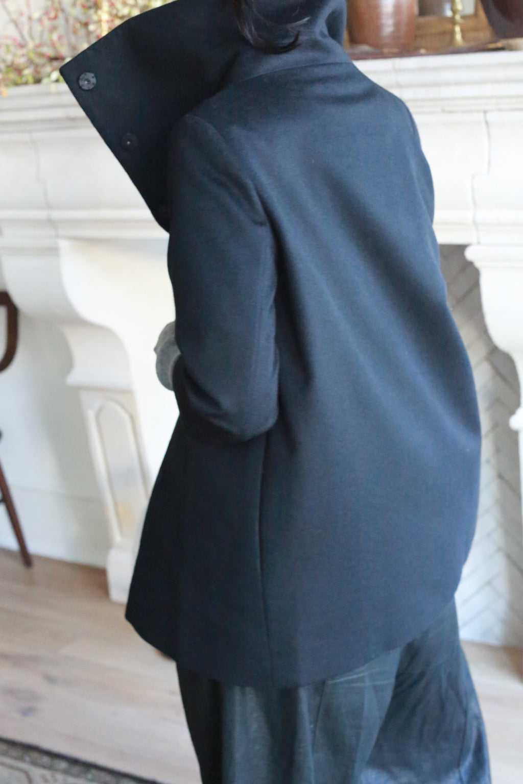 A person is standing with their back to the camera, wearing the Stamford coat in dark navy cashmere, featuring a high collar that obscures much of their face. They are in a room adorned with a light-colored fireplace and wooden furniture. Their posture appears relaxed.
