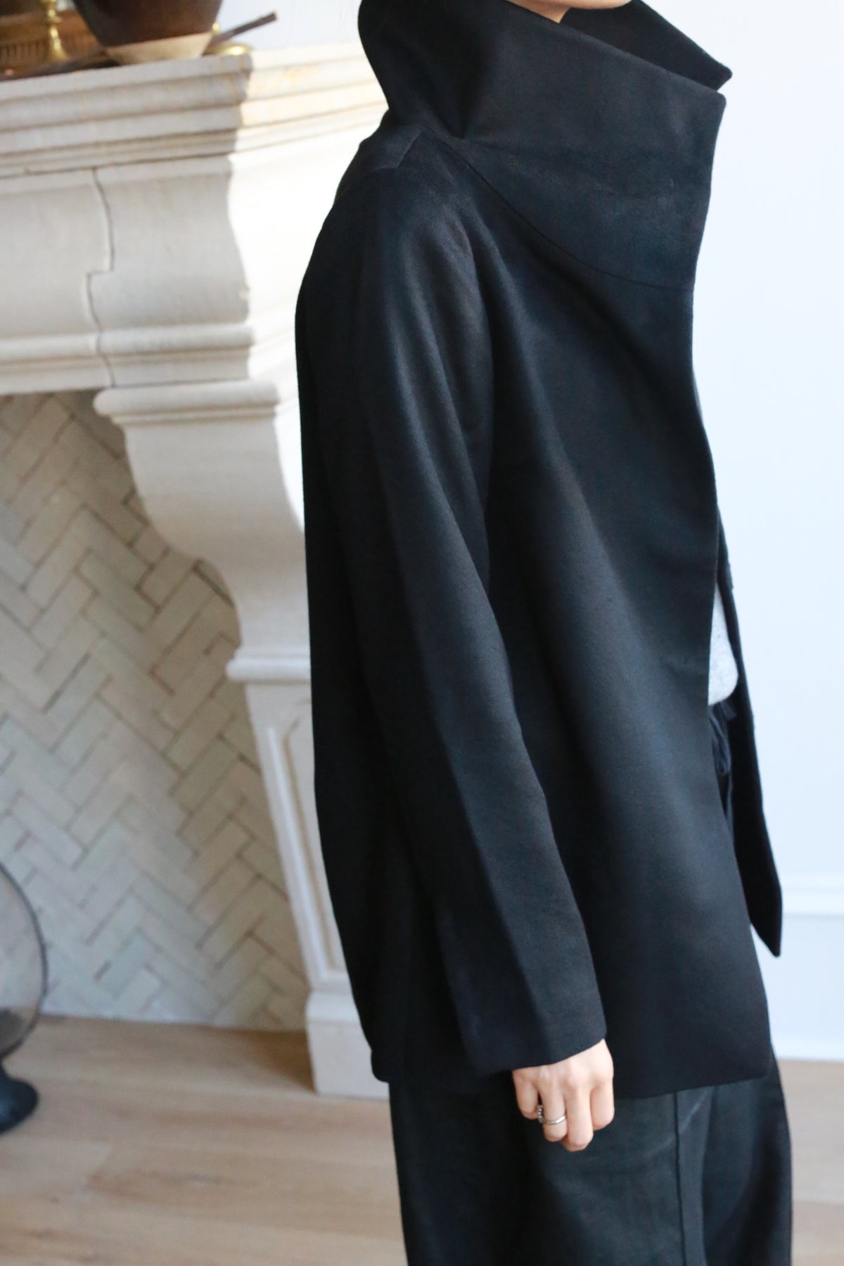 A person is standing indoors, partially visible from the shoulder down, wearing a Stamford II black high-collared jacket with their left hand at their side. The background features a light-colored, ornate fireplace with a herringbone-tiled wall and wooden floor.