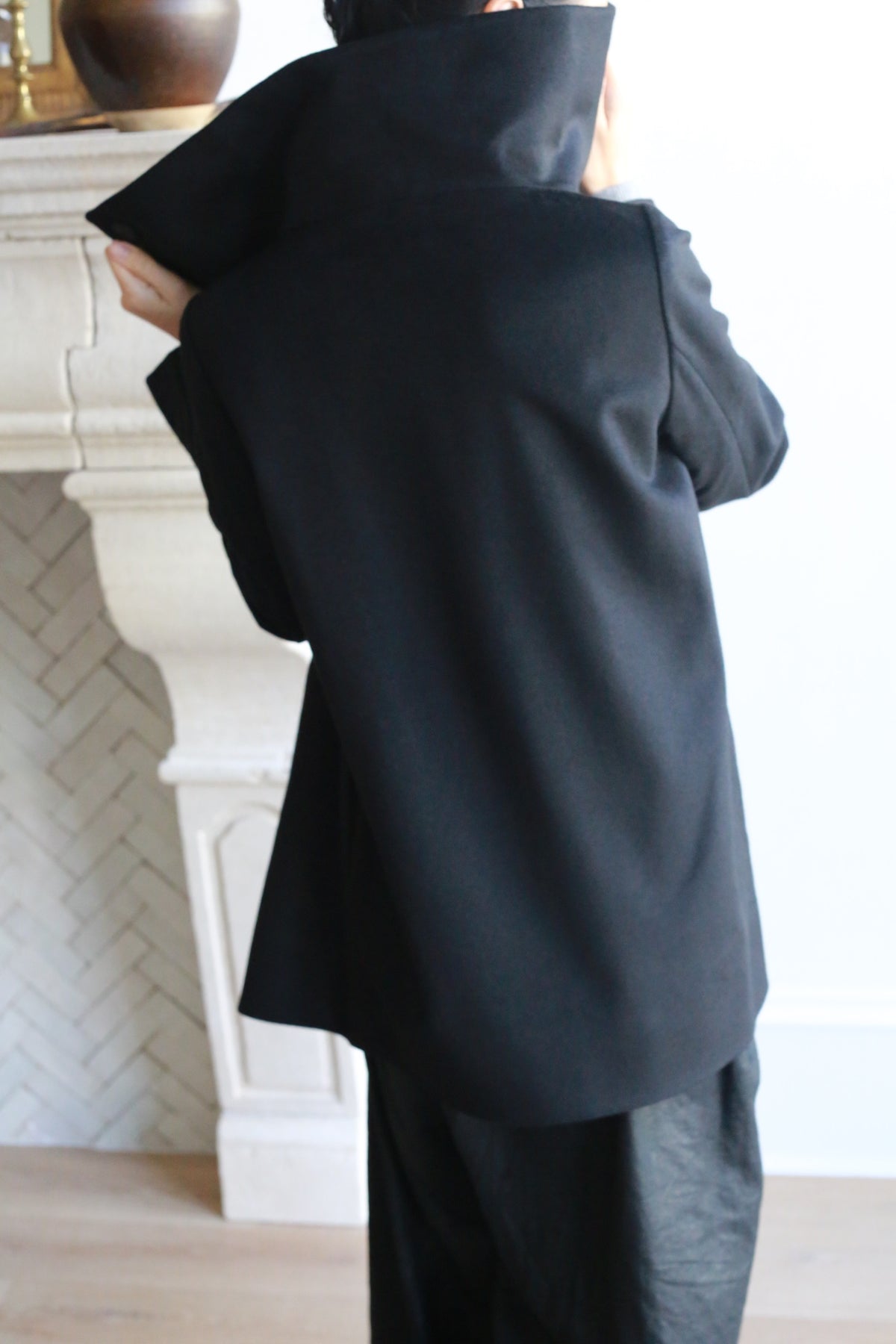A person is standing indoors with their back to the camera, dressed in a dark, oversized Stamford II jacket with the high collar turned up. The background features a section of a white, ornate fireplace and light-colored walls. The individual seems to be adjusting their Stamford II jacket with one hand.
