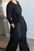 A charcoal gray short-sleeved cashmere Wexley | Black coat tunic with a tie waist and side pockets, worn over a long-sleeved black top and black wide-leg trousers.