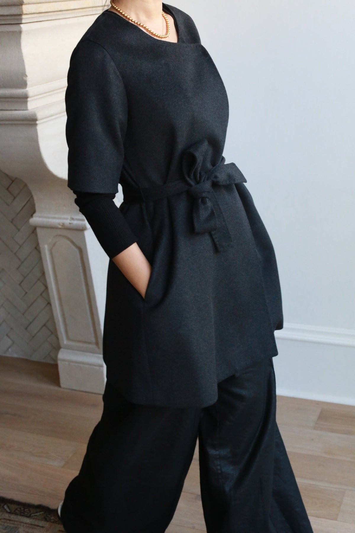 A charcoal gray short-sleeved cashmere Wexley | Black coat tunic with a tie waist and side pockets, worn over a long-sleeved black top and black wide-leg trousers.