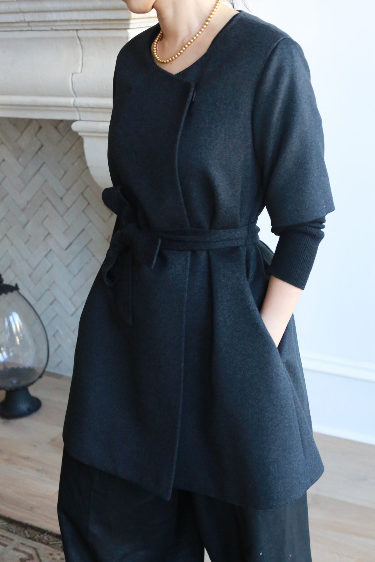 A person indoors, styled in the Wexley, a black woven cashmere coat with a three-quarter sleeve A-line cut and tied belt, paired with black pants. They accessorize with a gold necklace as they stand near a fireplace featuring herringbone design tiles in the background.