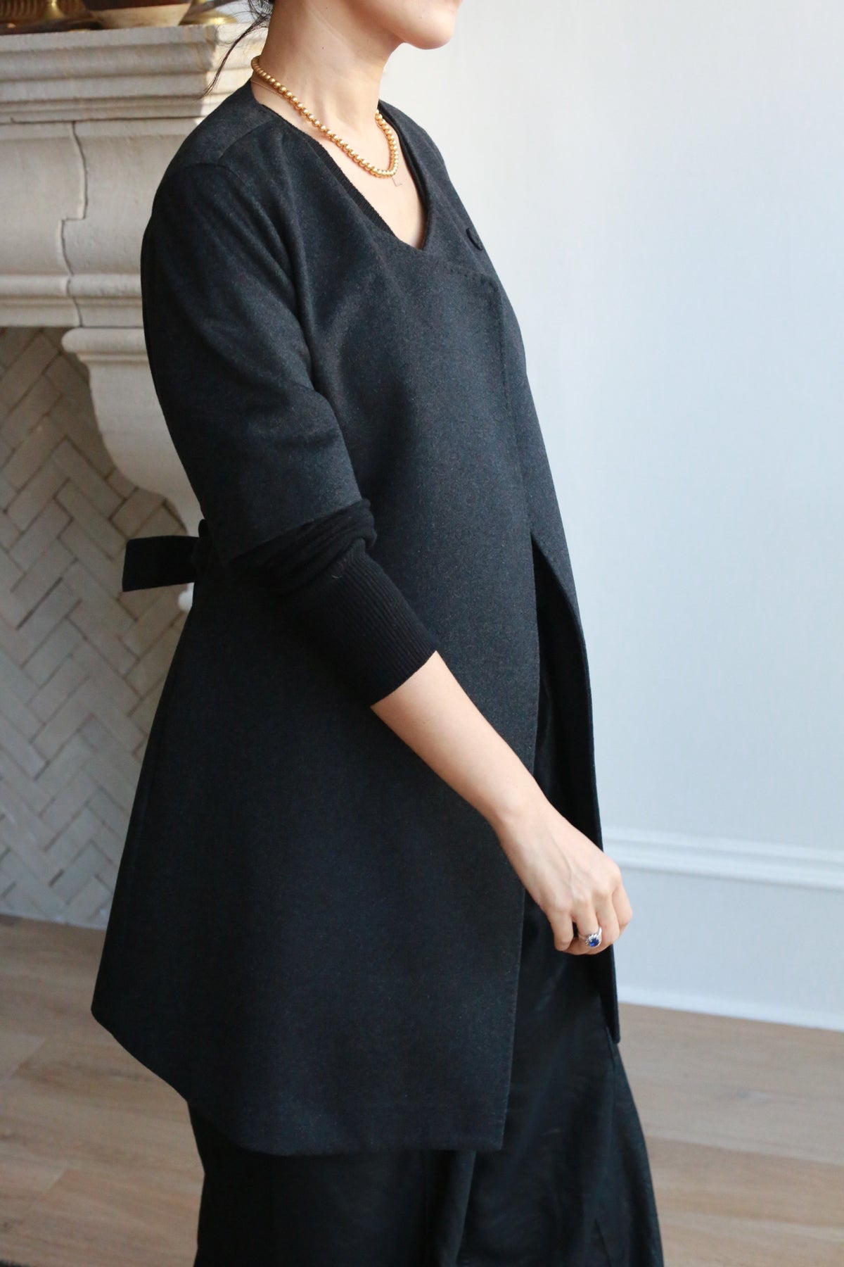 A person wearing the stylish Wexley black double-breasted coat and a gold necklace stands indoors near a white herringbone-patterned wall, hands loosely clasped, with a hint of a fireplace mantel visible in the background.