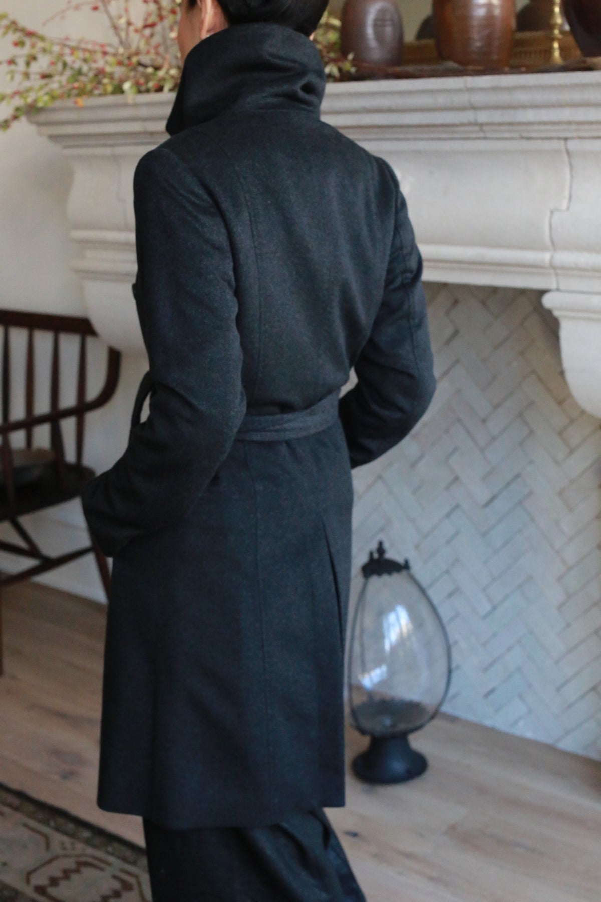A person in a dark Marylebone cashmere trench coat stands facing away in a cozy, elegant living room featuring a decorative fireplace, wooden chair, ornate floor vase, and light wooden flooring.