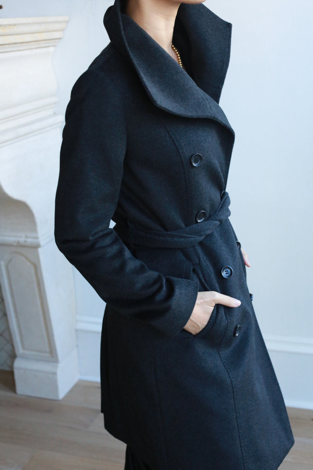 A person stylishly wearing the Marylebone double-breasted cashmere coat poses with hands in pockets, standing indoors by a white fireplace.