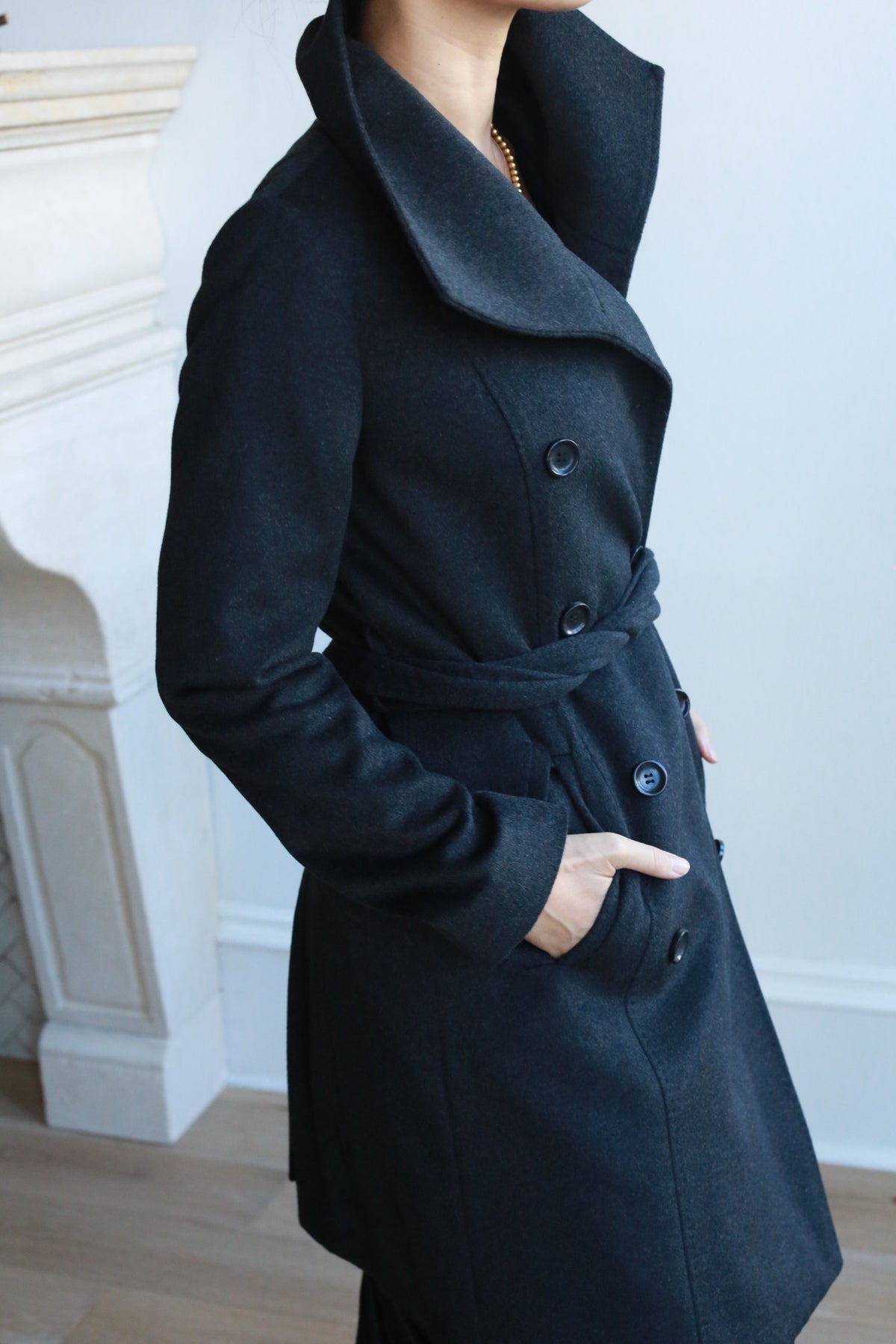 A person stylishly wearing the Marylebone double-breasted cashmere coat poses with hands in pockets, standing indoors by a white fireplace.