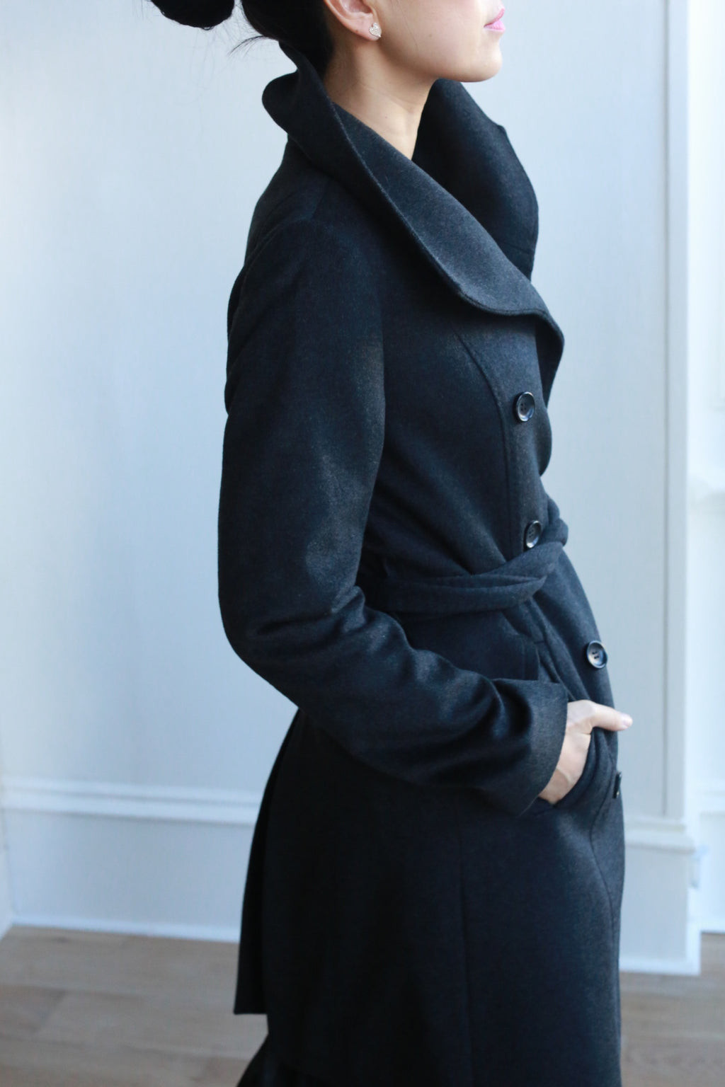 A person with dark hair in a bun wears the stylish Marylebone black cashmere coat, featuring a high collar and tied belt. The double-breasted design shines as they pose sideways with one hand in a pocket against a softly lit white wall.