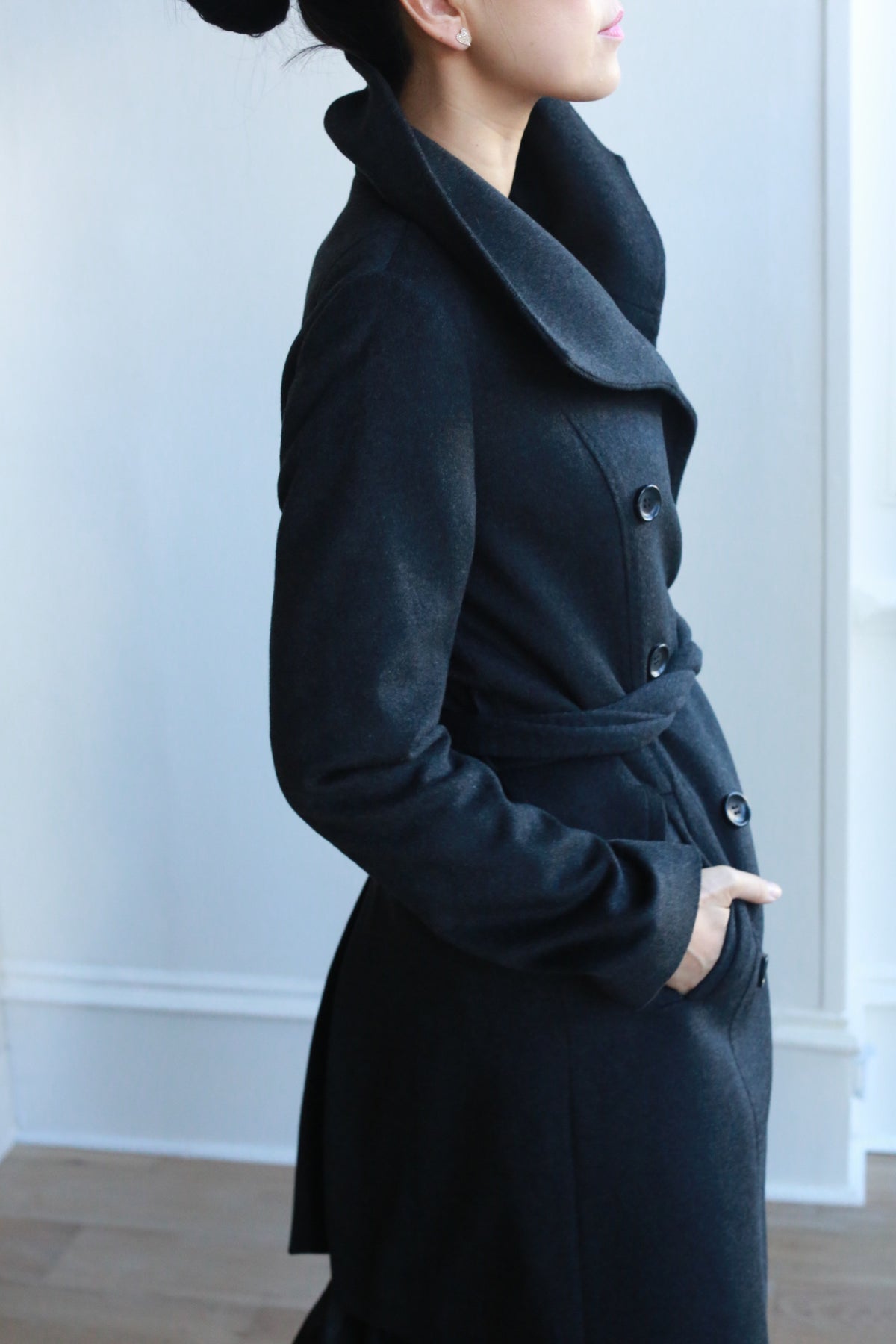 A person with dark hair in a bun wears the stylish Marylebone black cashmere coat, featuring a high collar and tied belt. The double-breasted design shines as they pose sideways with one hand in a pocket against a softly lit white wall.