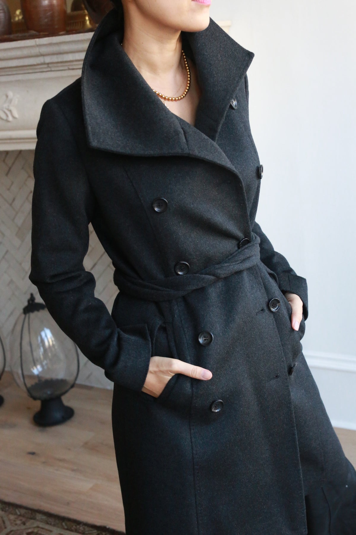 Wearing the Marylebone trench coat—a dark, double-breasted piece with a high collar, belt, and cashmere blend—a person stands with one hand in their pocket against a backdrop of a fireplace and decorative elements.
