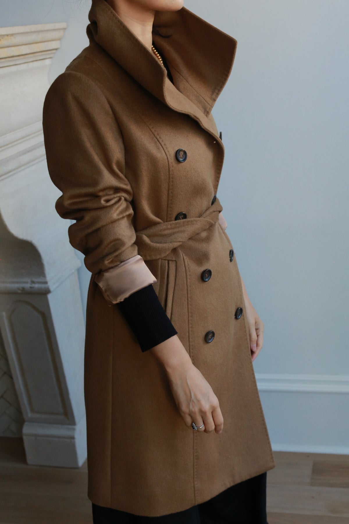 Indoors, a person wears the elegant Marylebone double-breasted trench coat in stylish brown cashmere with dark buttons, a belted waist, and a high collar. In the background is part of a fireplace against a light-colored wall.