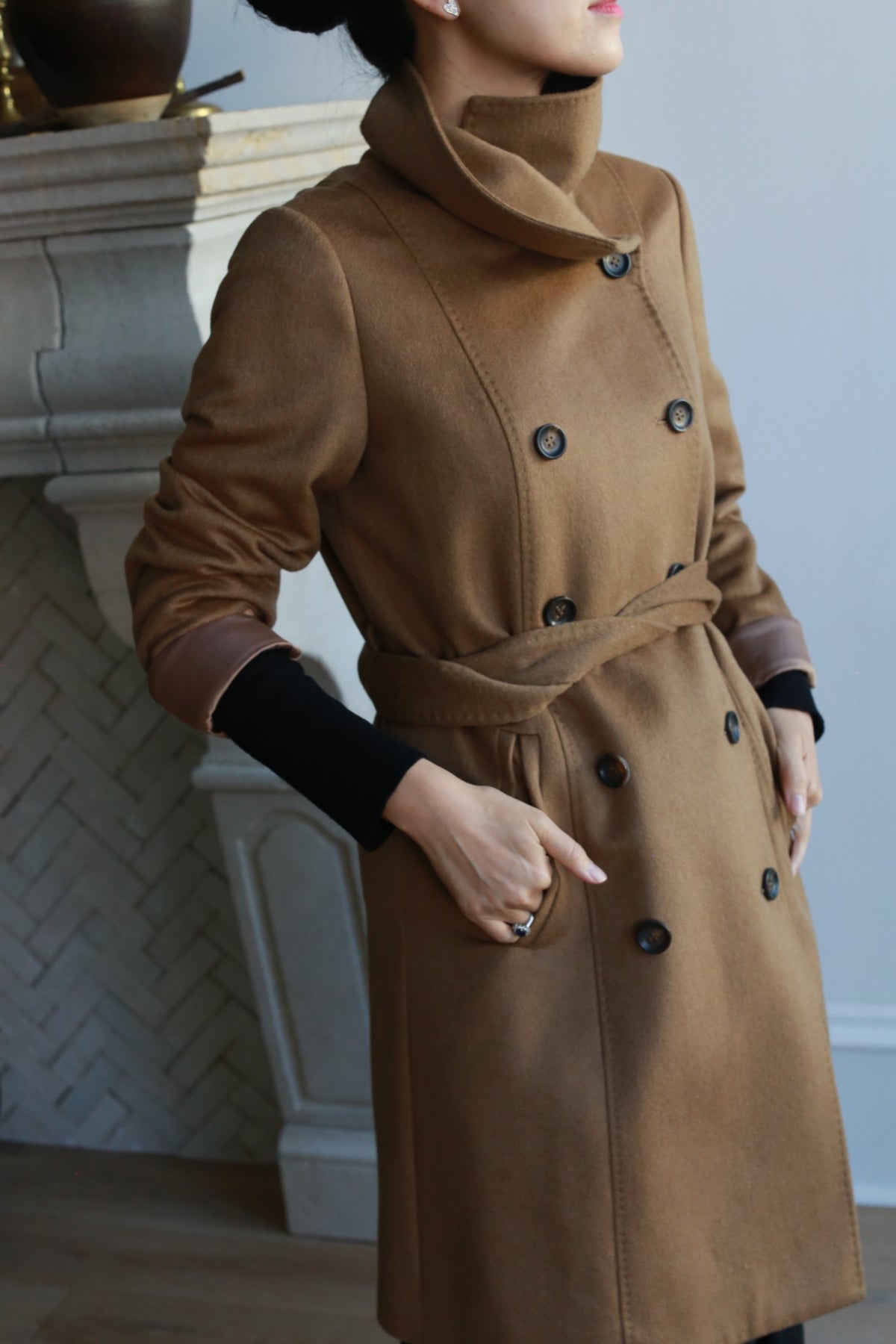 A person in a Marylebone cashmere double-breasted coat with a high collar and belt stands with hands in pockets by a white brick fireplace.