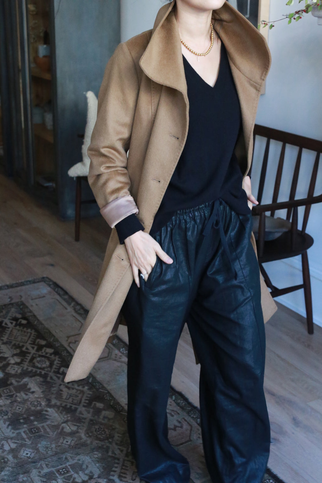Indoors, a person wears the Marylebone, a double-breasted tan coat over a black V-neck top and black pants. Their right hand rests in the coat pocket, accompanied by a gold necklace. A wooden chair and patterned rug are visible in the background.