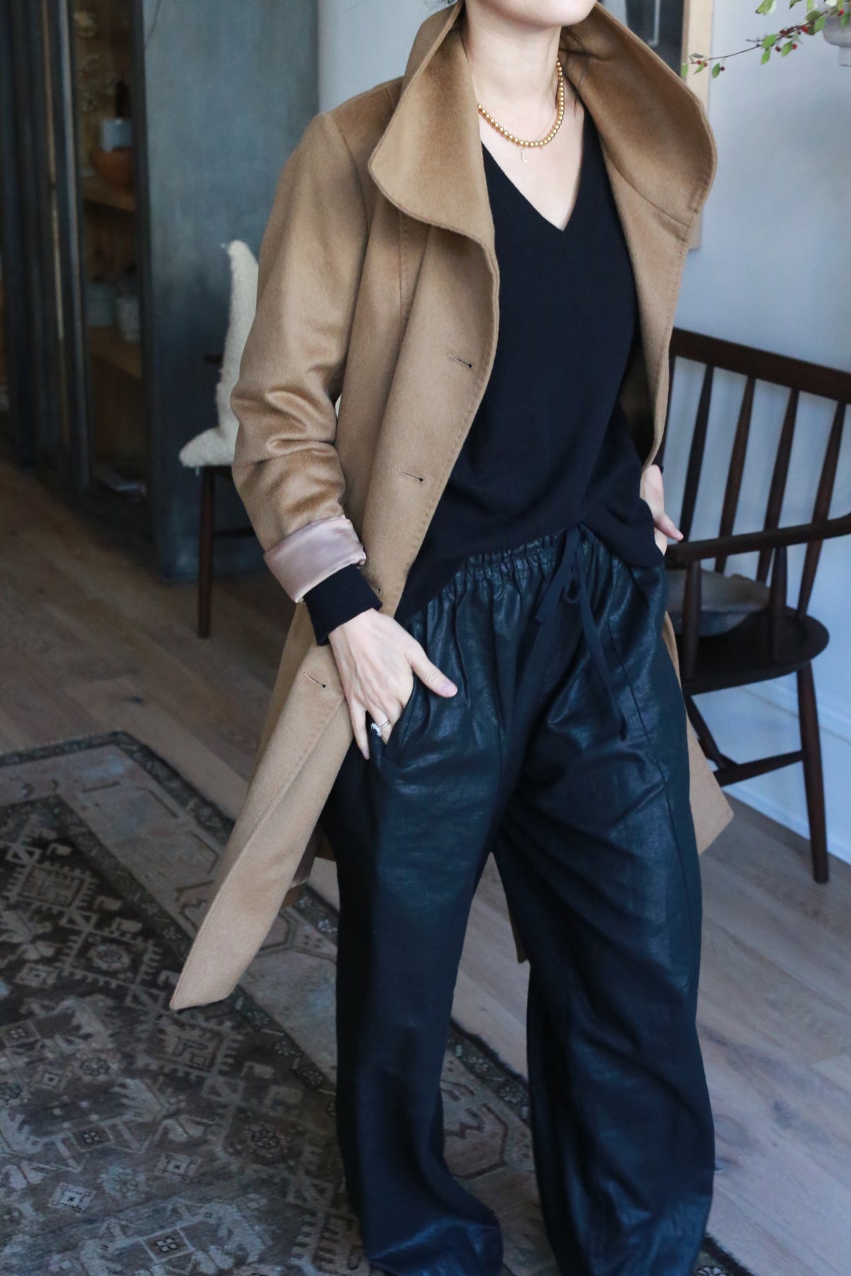 Indoors, a person wears the Marylebone, a double-breasted tan coat over a black V-neck top and black pants. Their right hand rests in the coat pocket, accompanied by a gold necklace. A wooden chair and patterned rug are visible in the background.