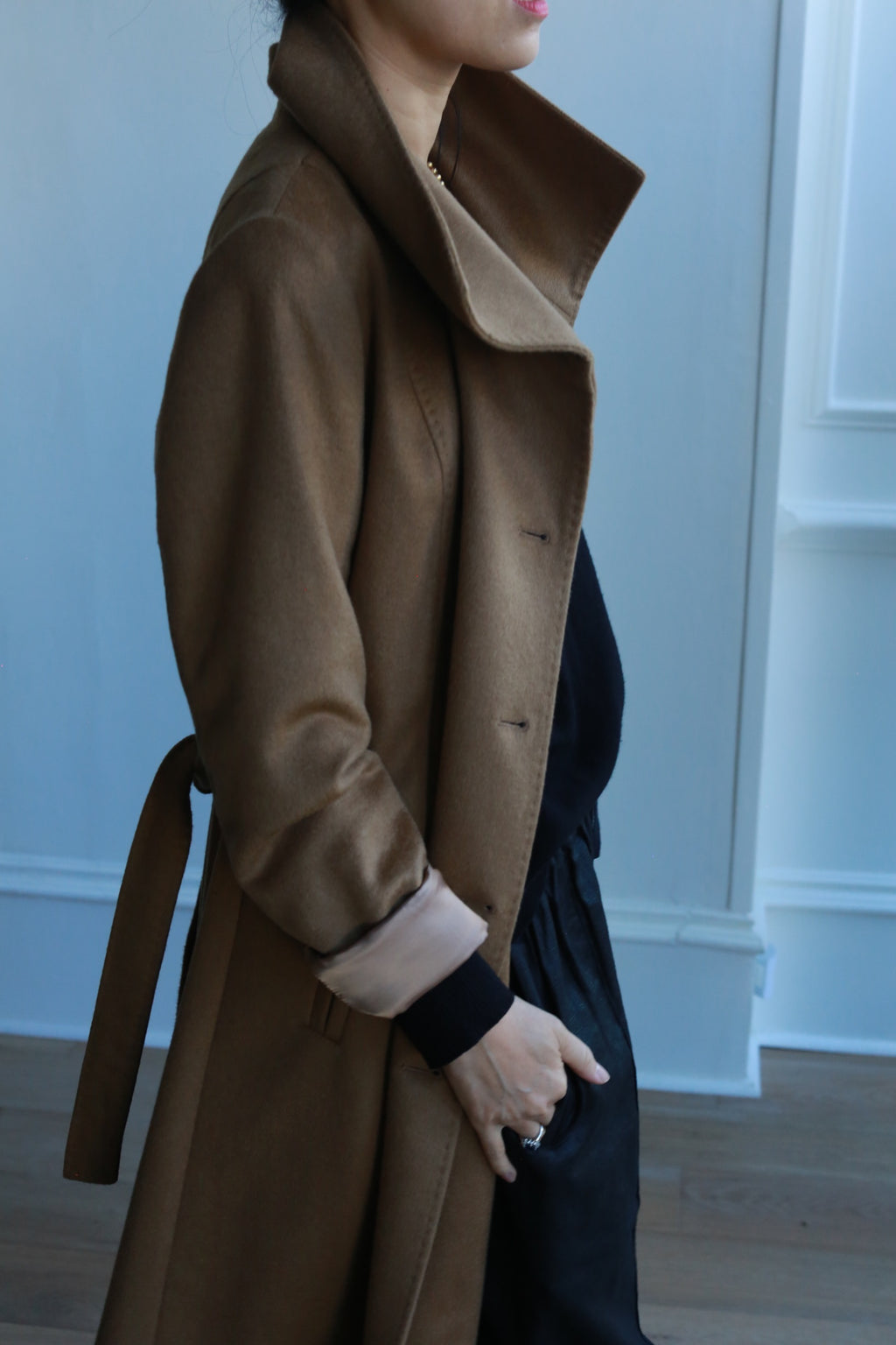 A person in a Marylebone brown trench coat with a high collar stands in a room with white walls and wooden flooring. The double-breasted coat features a belt, and the individual is wearing dark pants and a black top. The image is partially cropped, revealing the upper body.