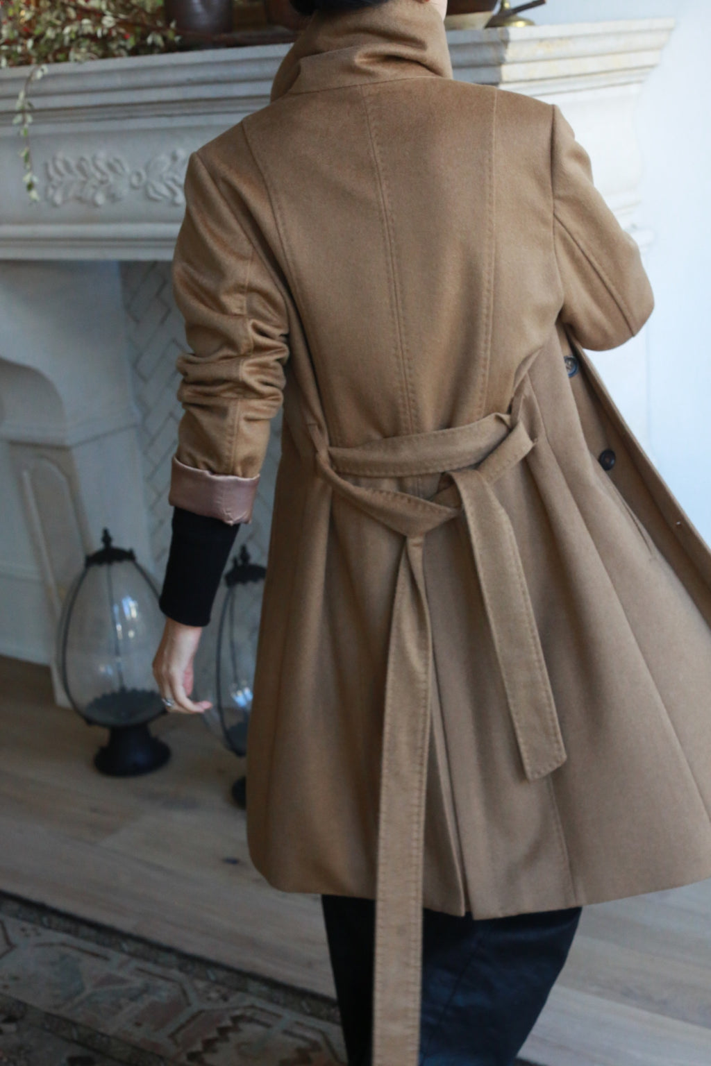 In a room with wooden floors and a white fireplace, someone wears the Marylebone, a long double-breasted trench coat with a belt, while two decorative lanterns enhance the cozy ambiance.