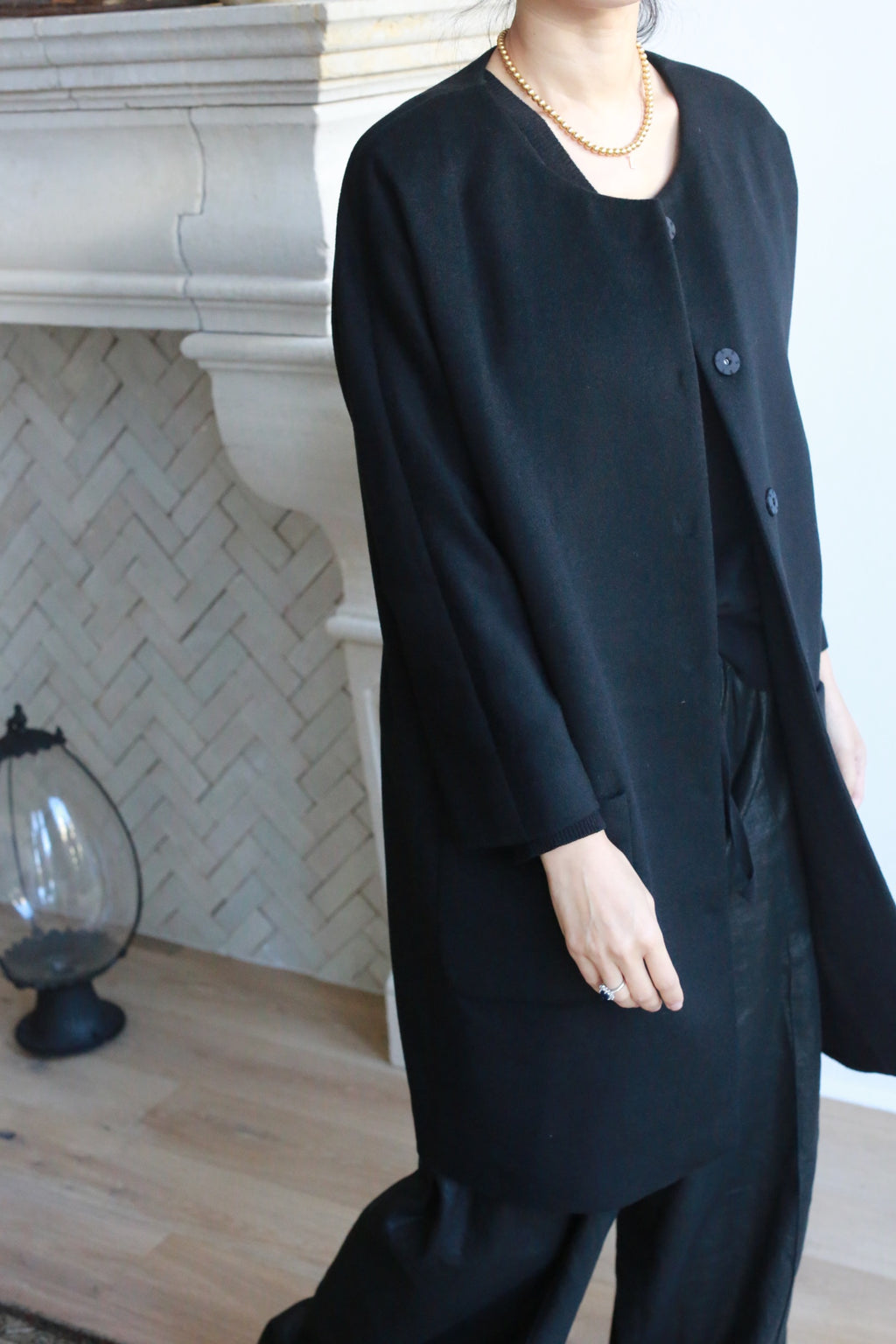 Someone wears the Battersea long black cashmere coat with handmade snap buttons, paired with black pants and a gold necklace. They stand indoors near a decorative fireplace, their face partially out of frame.