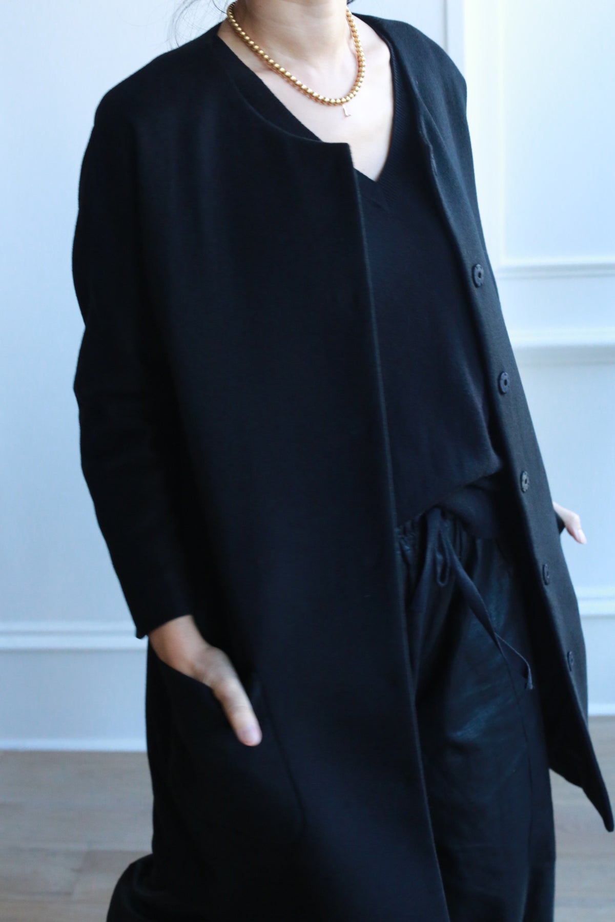 Wearing the Battersea black cashmere coat with raglan sleeves, a person stands indoors against a white wall, styled with a black top and pants, a gold beaded necklace, and one hand in their pocket.
