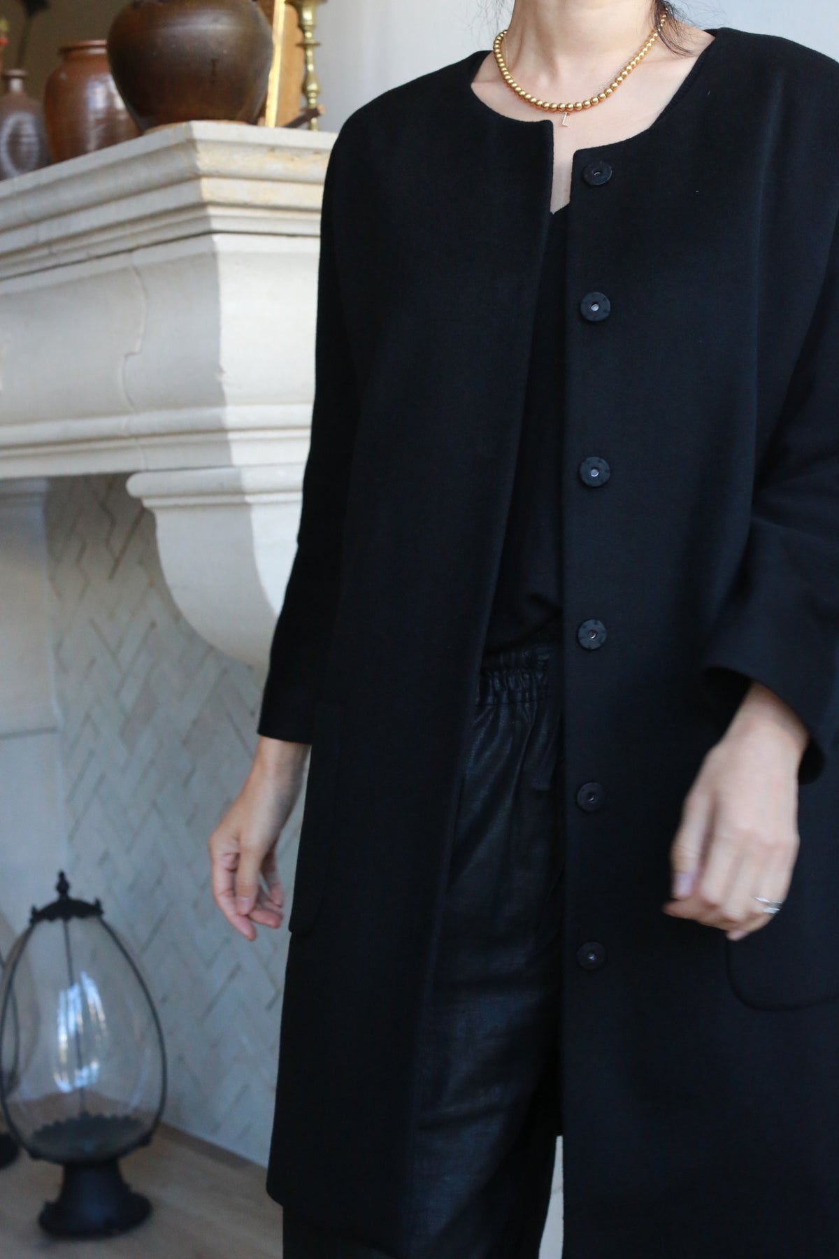 A person stands indoors near a white fireplace, wearing the Battersea long black cashmere coat with handmade snap buttons, paired with a gold necklace, black shirt, and black pants. The image is cropped from the shoulders down.