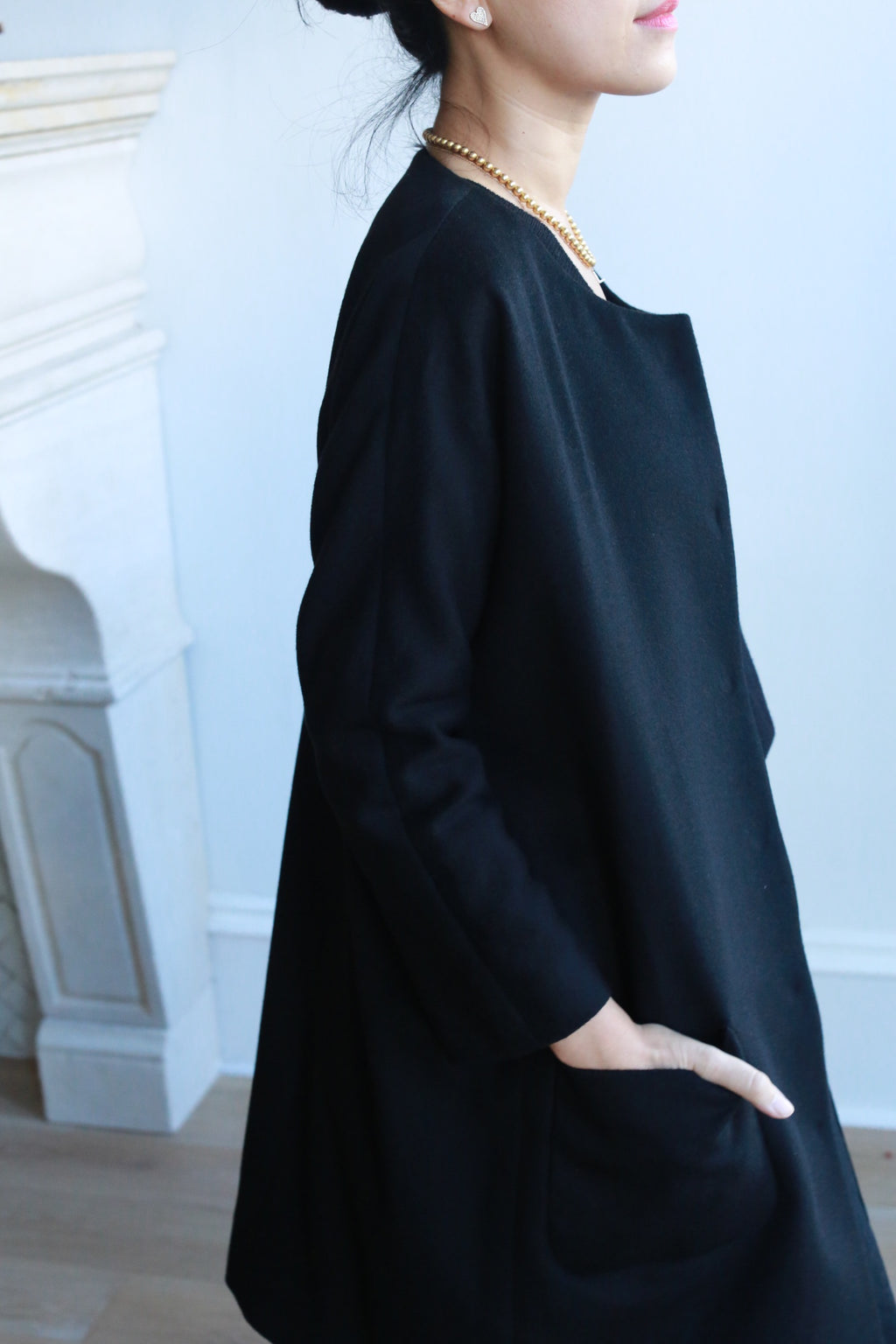 A woman stands near a white fireplace with one hand in her pocket, wearing the Battersea black long-sleeve dress with raglan sleeves and a pearl necklace. Her hair is tied back, and the photo is cropped to show her upper body and part of her face.