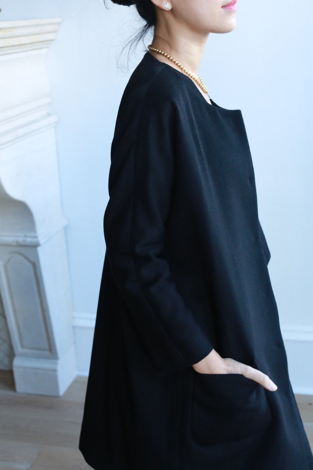 A woman stands near a white fireplace with one hand in her pocket, wearing the Battersea black long-sleeve dress with raglan sleeves and a pearl necklace. Her hair is tied back, and the photo is cropped to show her upper body and part of her face.